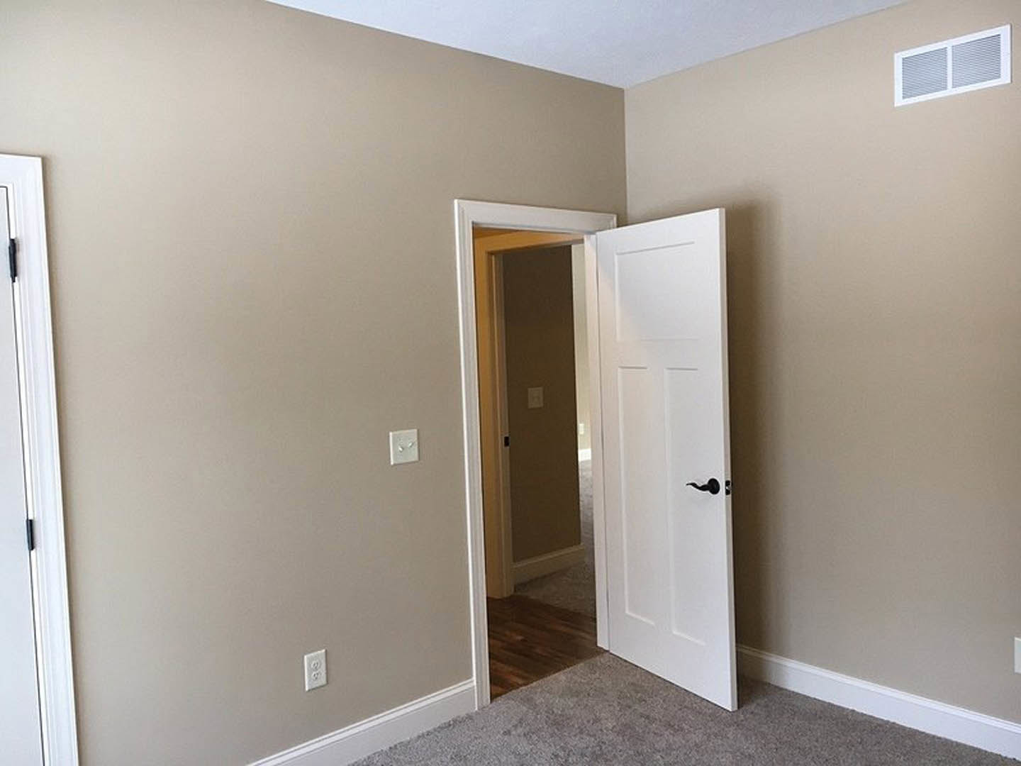 White door with silver handle open in a room, wood floor beneath, white light switch and vent visible on plaster wall