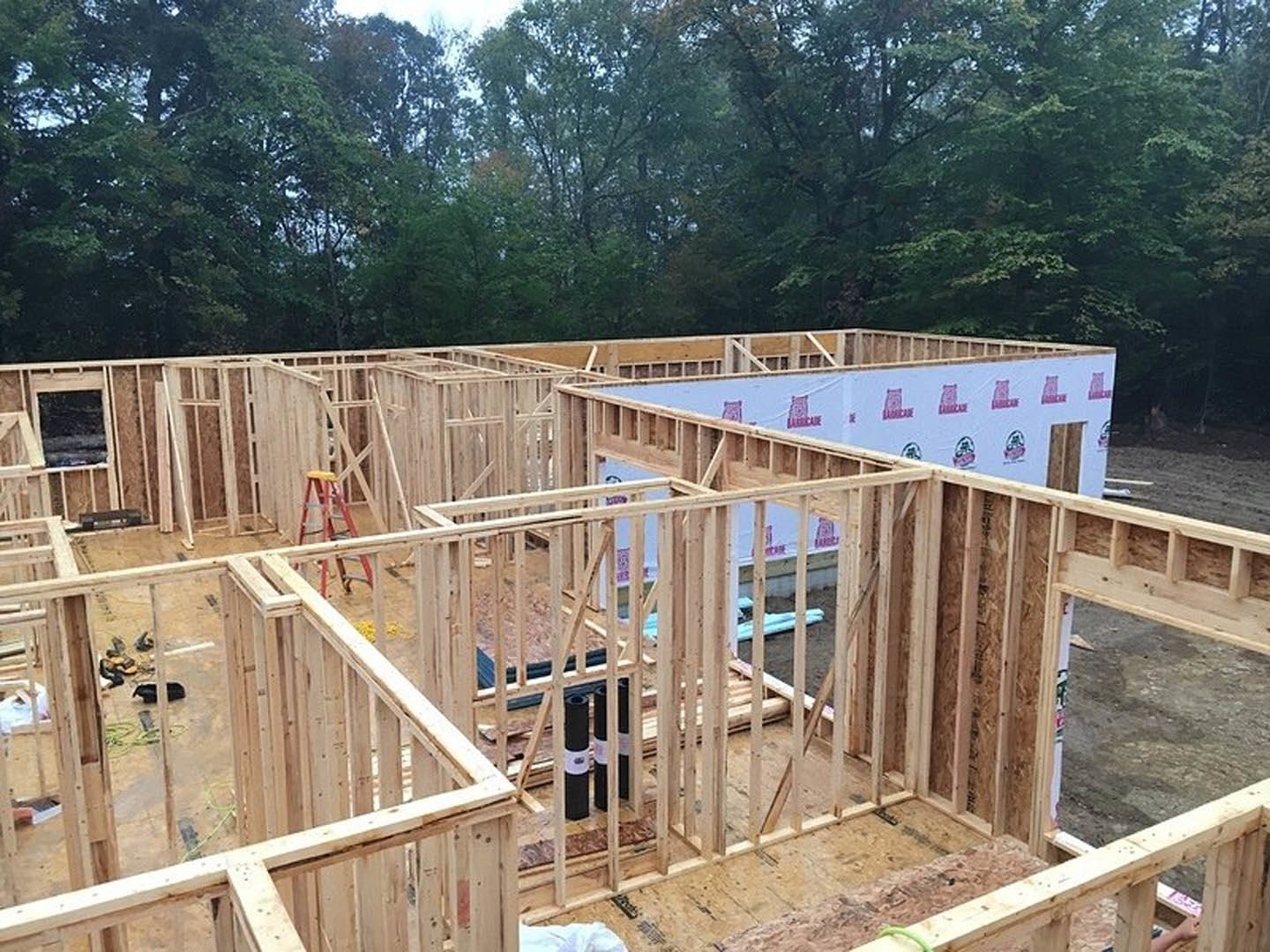 Partially constructed wooden house with exposed framing, ladder leaning against exterior wall, trees and fence in background, construction sign attached to wall