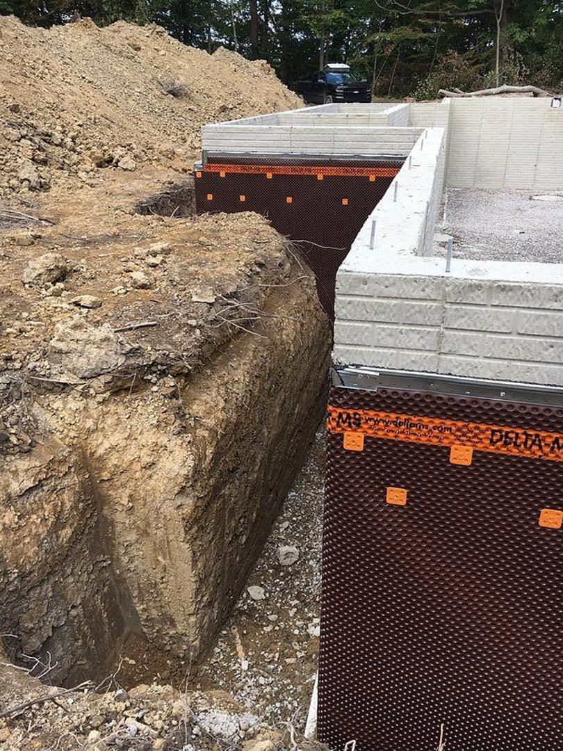 Excavated construction site with exposed foundation, large dirt mound, brown and orange soil, parked car, and scattered rocks and trees in the background