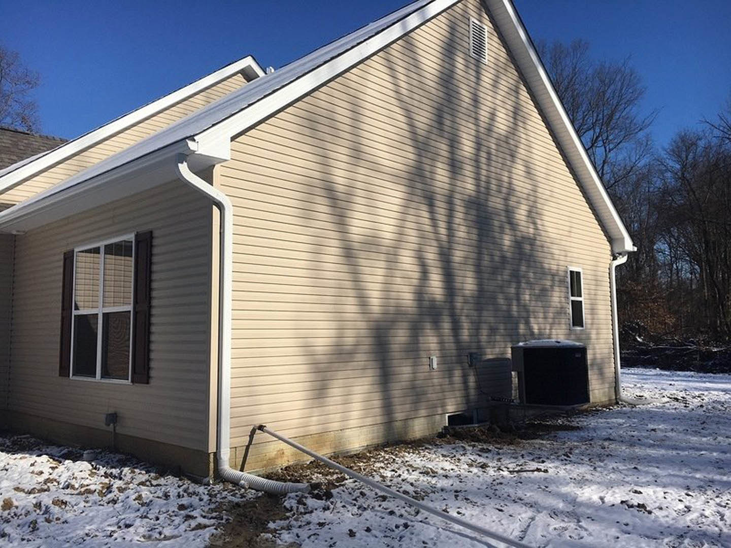Two-story home with light siding, large windows with blinds, tree shadow cast on snowy yard, white pipe visible in snow near exterior wall.