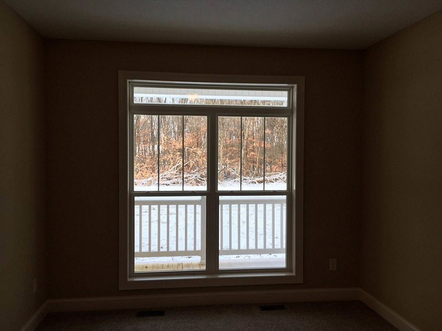 Carpeted room with large window overlooking snowy forest, deck visible through glass, white walls, porch railing in foreground