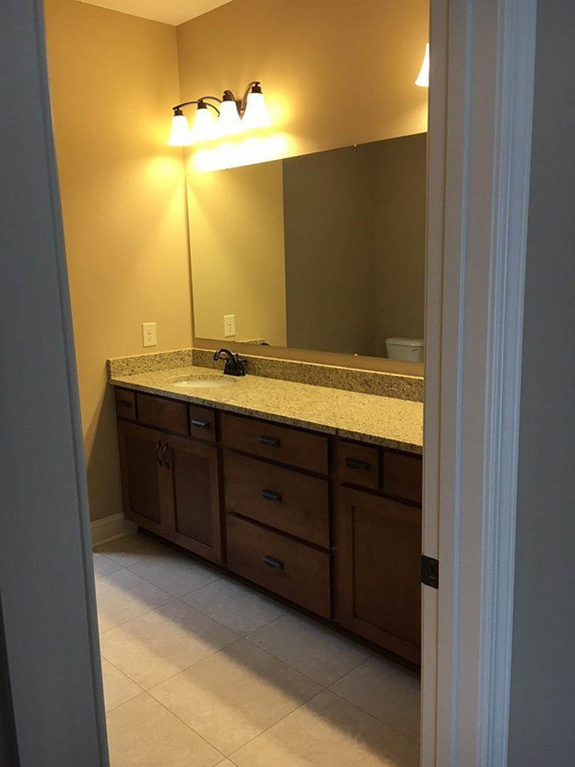 Bathroom with marble countertop, large wall mirror framed by mounted lights, tile flooring, and built-in cabinetry with drawers.