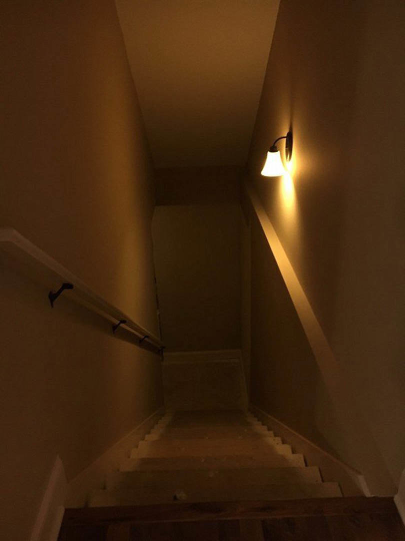 Wood staircase with white plaster walls, recessed ceiling light, and modern wall sconce illuminating the steps.