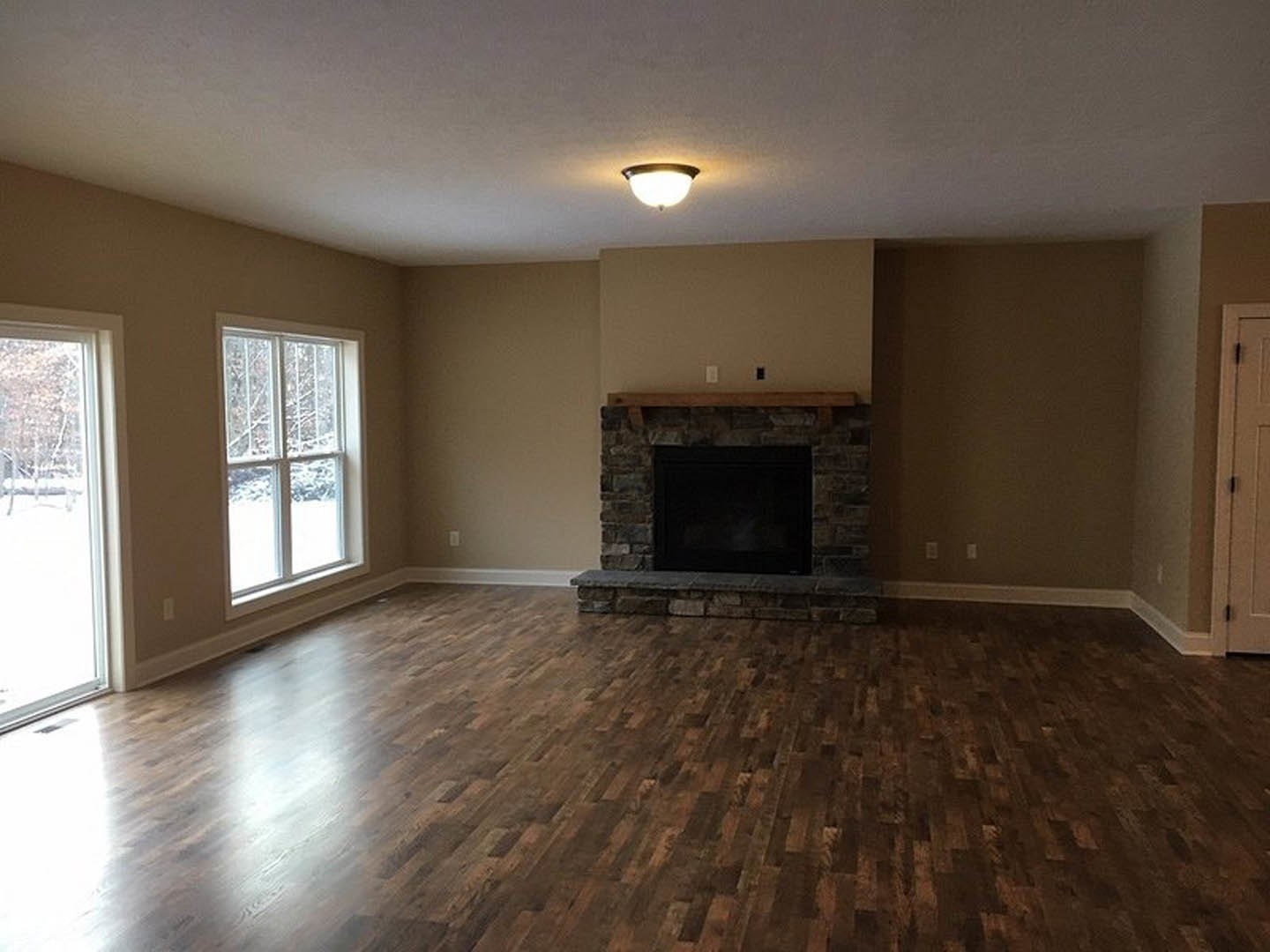 Living room with hardwood flooring, white plaster walls, black-framed fireplace with glass screen, wall-mounted light fixture, large window showing snowy landscape