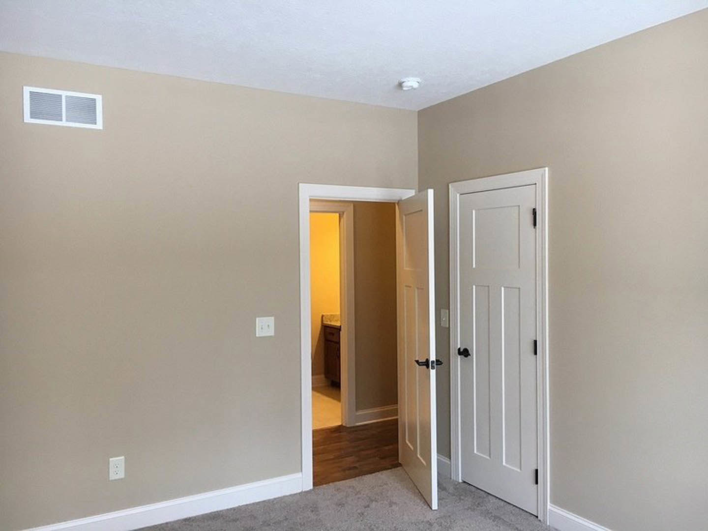 Open white door with brushed metal handle, wood plank flooring, wall vent, and doorway leading to tiled bathroom