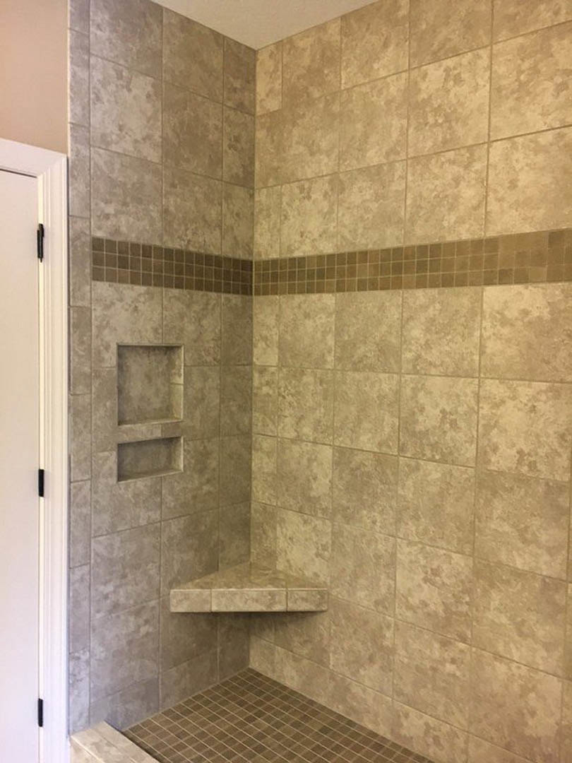Walk-in shower featuring a built-in shelf, white plaster walls with black hardware, brown tile flooring with white grout, and stone and marble accent walls