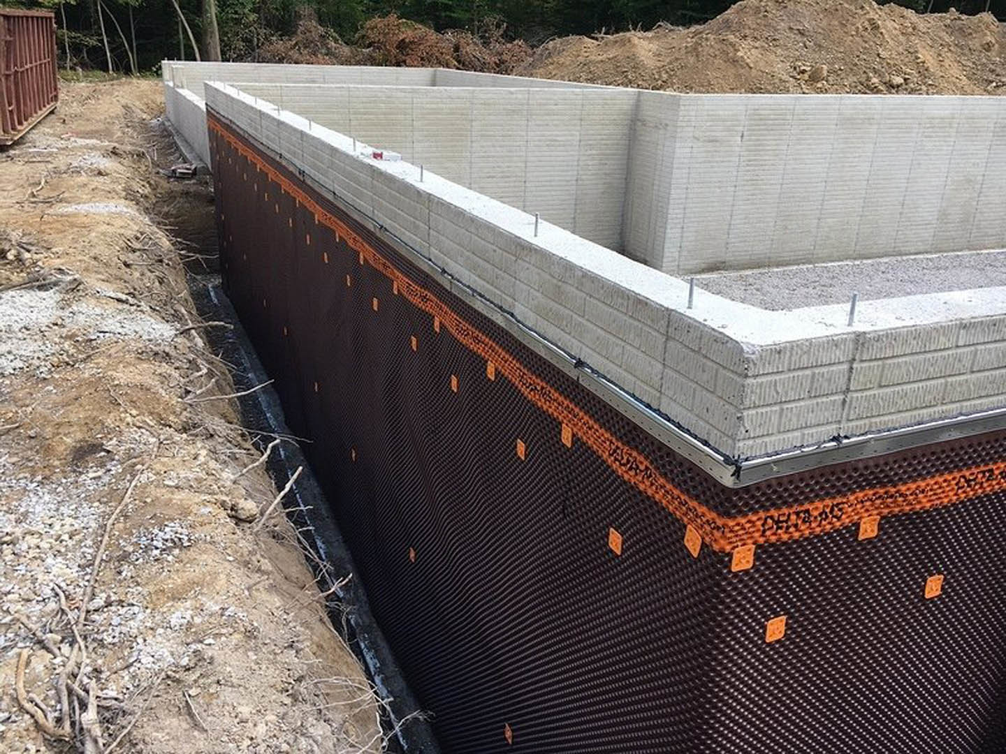 Concrete foundation slab with embedded metal rebar, surrounded by dirt and scattered rocks, outdoor construction site with blurred trees in background