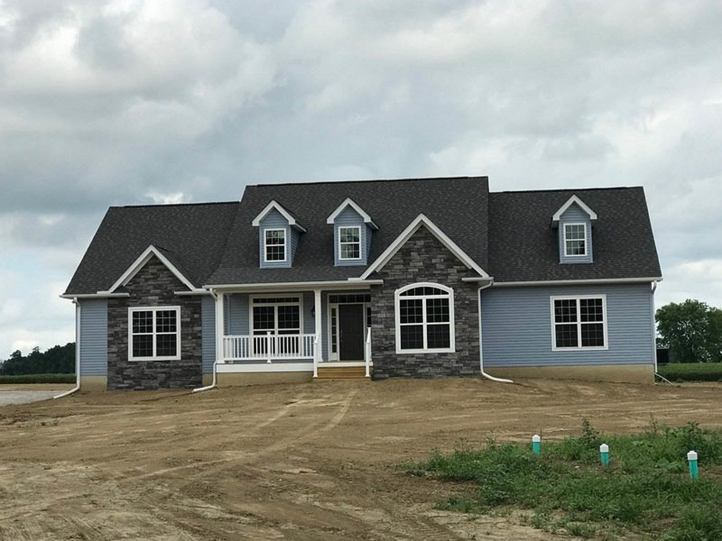 Two-story farmhouse with white railing and porch, dirt yard in foreground, green poles near window, leafy tree beside house, cloudy sky overhead