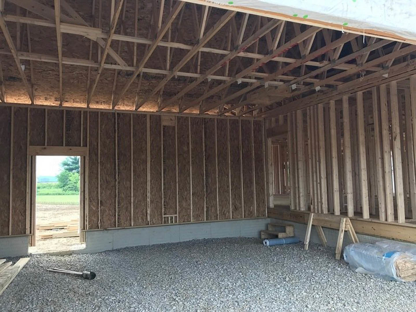 Wood plank ceiling and exposed beams above a gravel floor, window overlooking field and trees, plastic-wrapped object near wall.