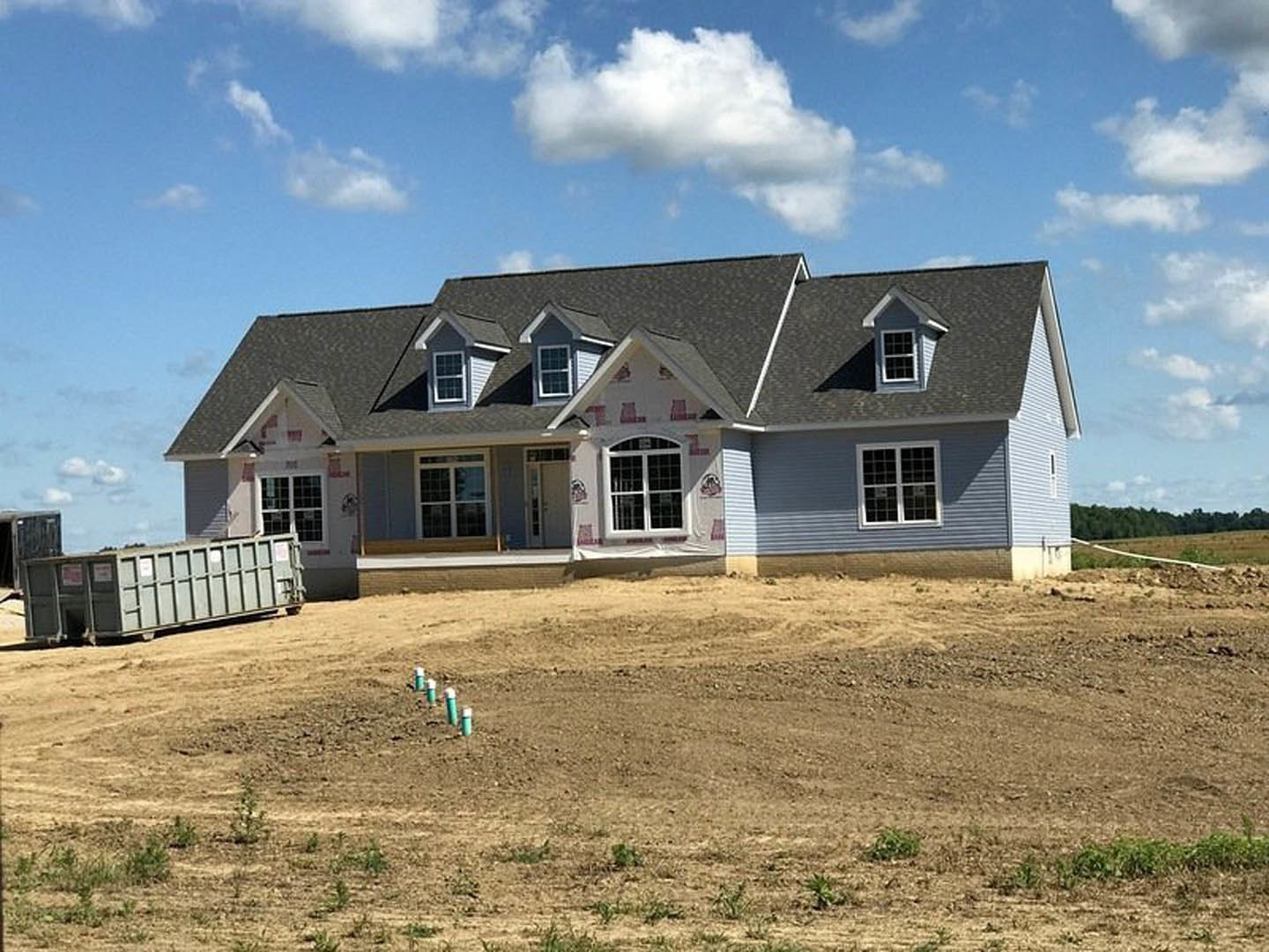 Modern house with grey roof and white-framed window, surrounded by a large dirt field with scattered utility poles; grey shipping container on a truck parked nearby under a partly