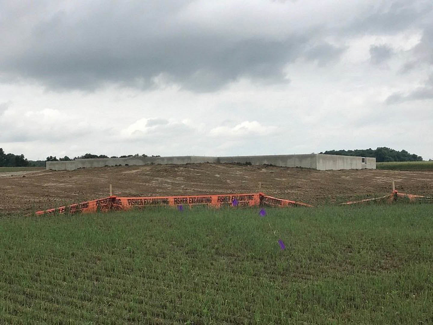 Wide grassy field bordered by a wooden fence, brick building with construction sign in background, overcast sky with dense clouds