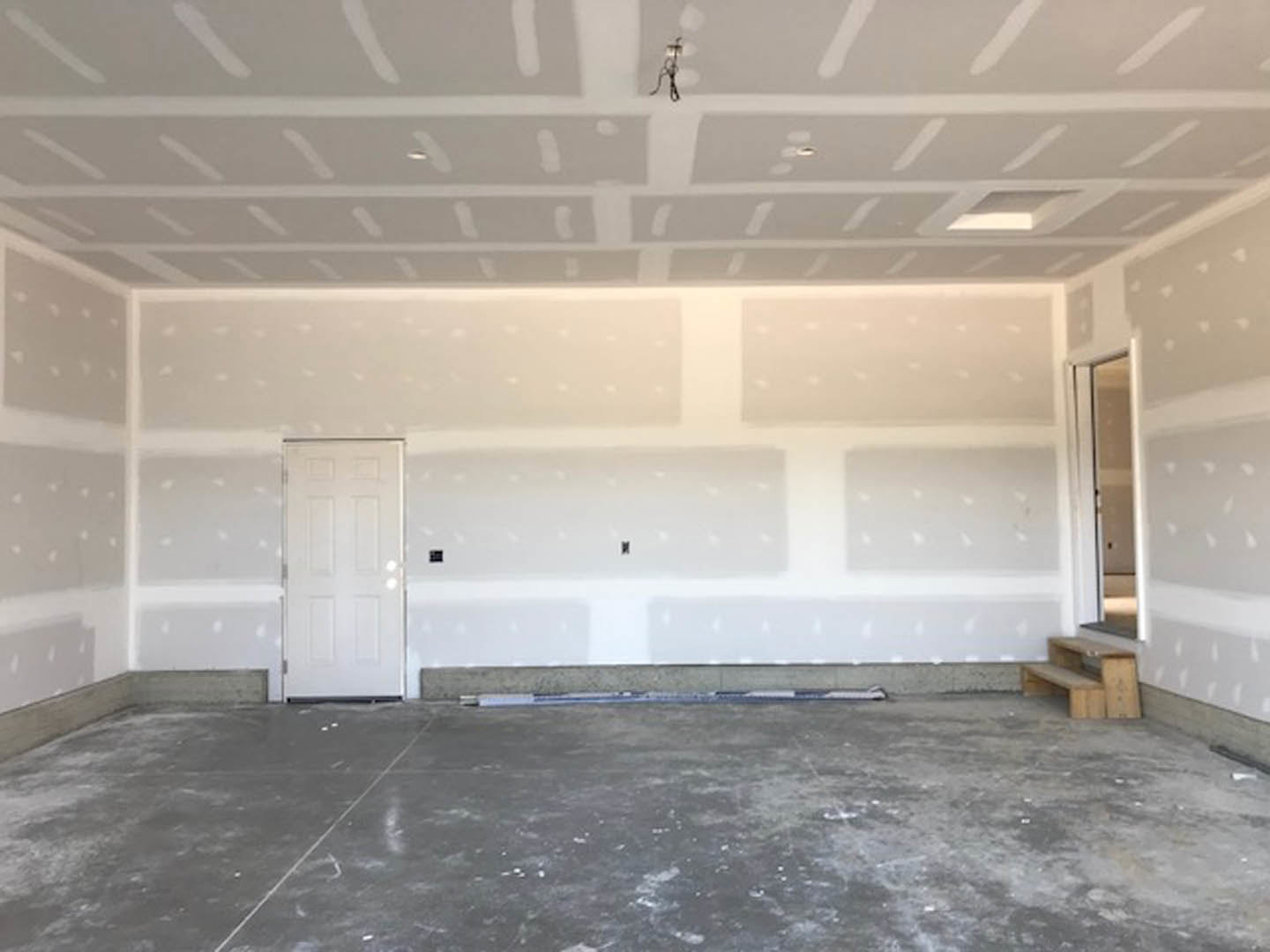 Open room with white plaster walls, wooden staircase, concrete floor, white door, and ceiling-mounted light fixture
