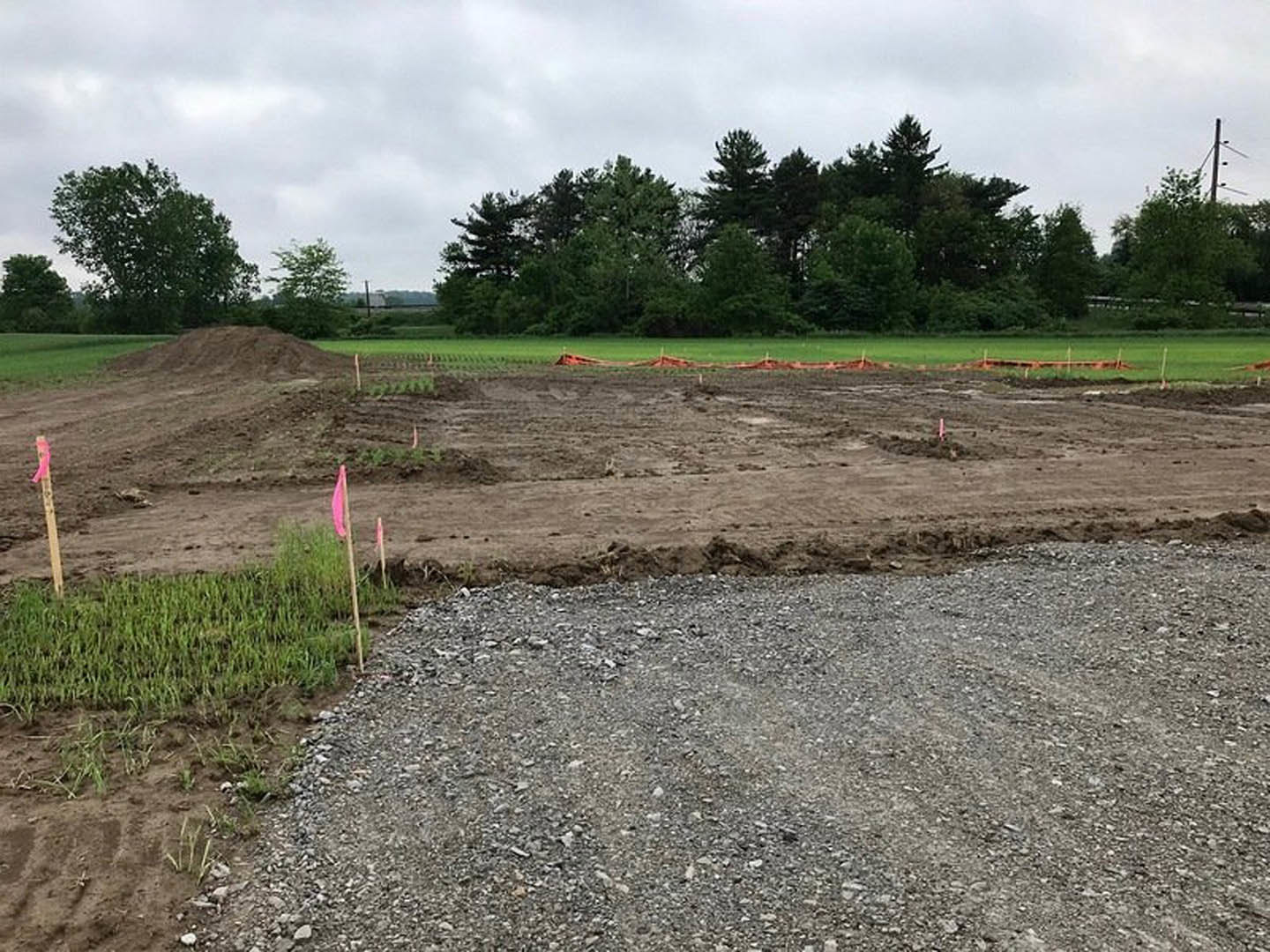 Dirt field marked with orange flags, gravel road in foreground, wooden post with holes, group of trees under cloudy sky