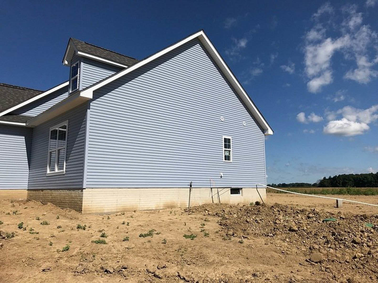 Partially built house with exposed framing and unfinished exterior, set in a grassy dirt field under a blue sky with scattered clouds