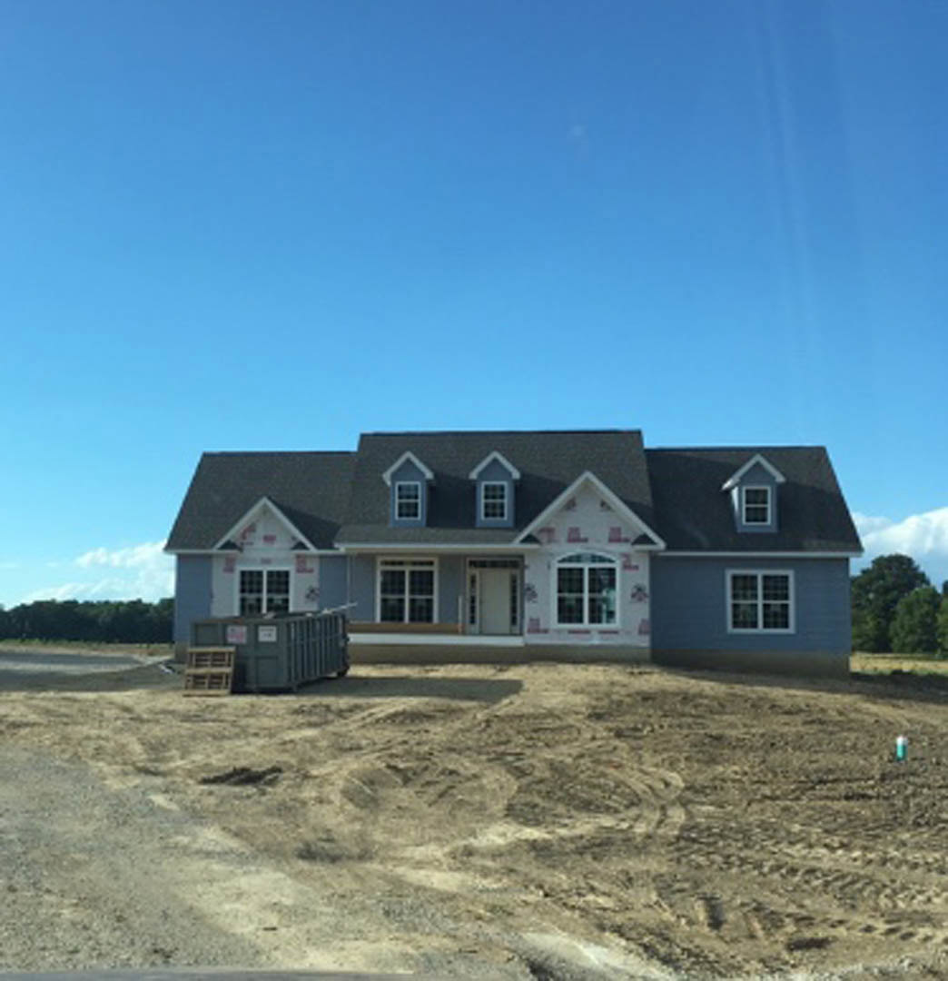 Blue-roofed house with white-framed window, large metal container displaying signs, dirt field marked by tire tracks, sparse trees, and overcast sky