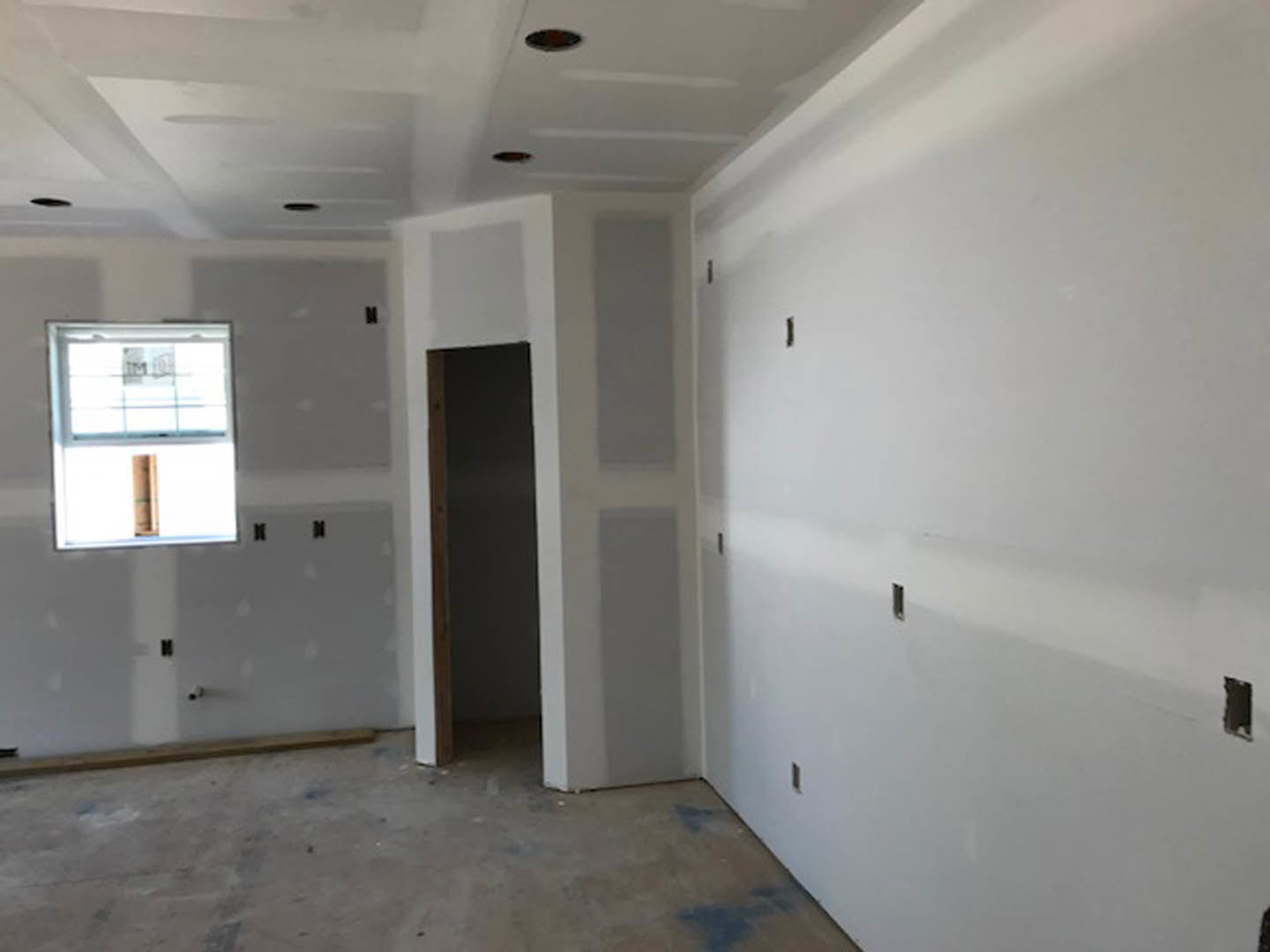 White plaster walls, unfinished concrete floor, window partially covered with cardboard, door visible, ceiling and wall fixtures not yet installed