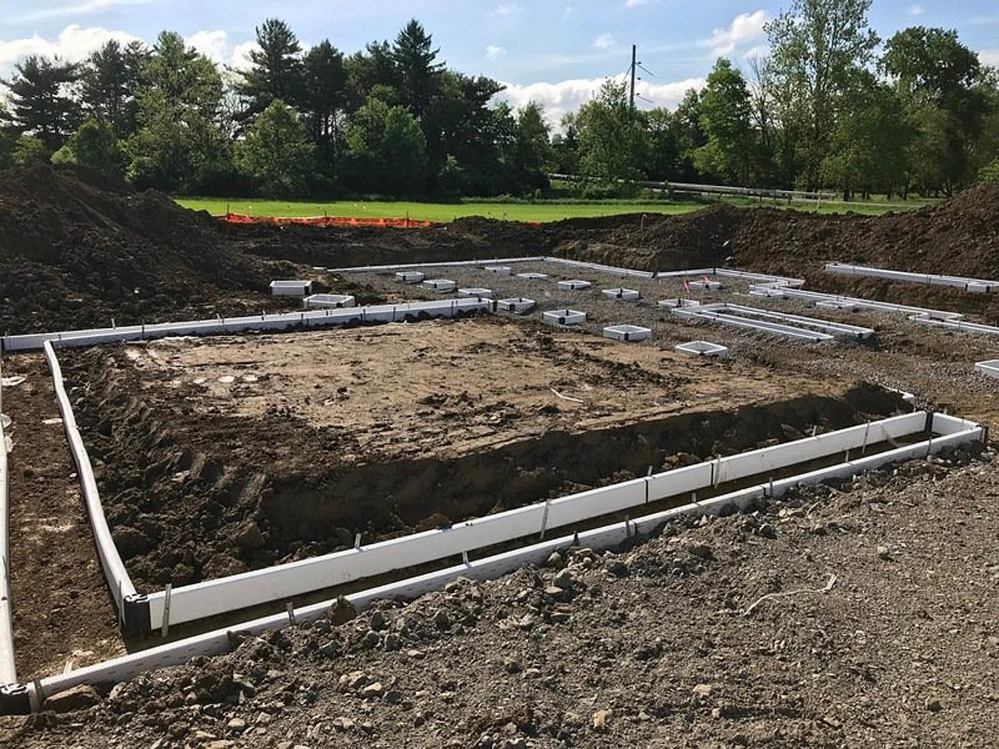 White foundation poles and a rectangular board set in dirt at a residential construction site, surrounded by trees and open sky.