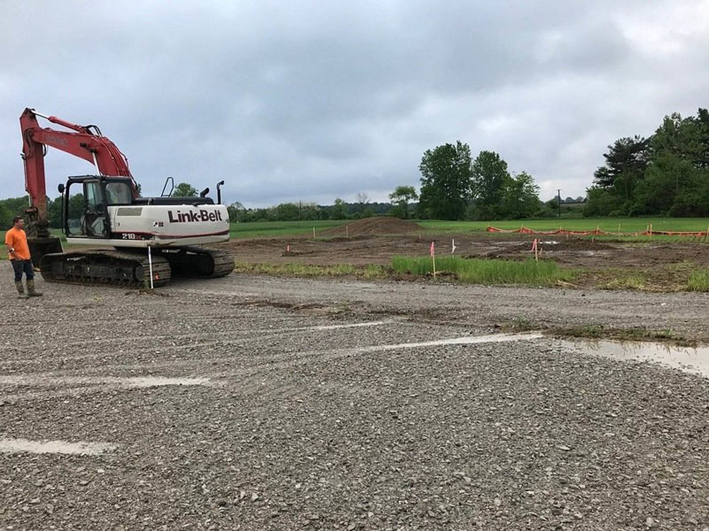 Bulldozer with crane attachment parked on dirt road near grassy field, man in orange shirt standing beside vehicle, overcast sky overhead