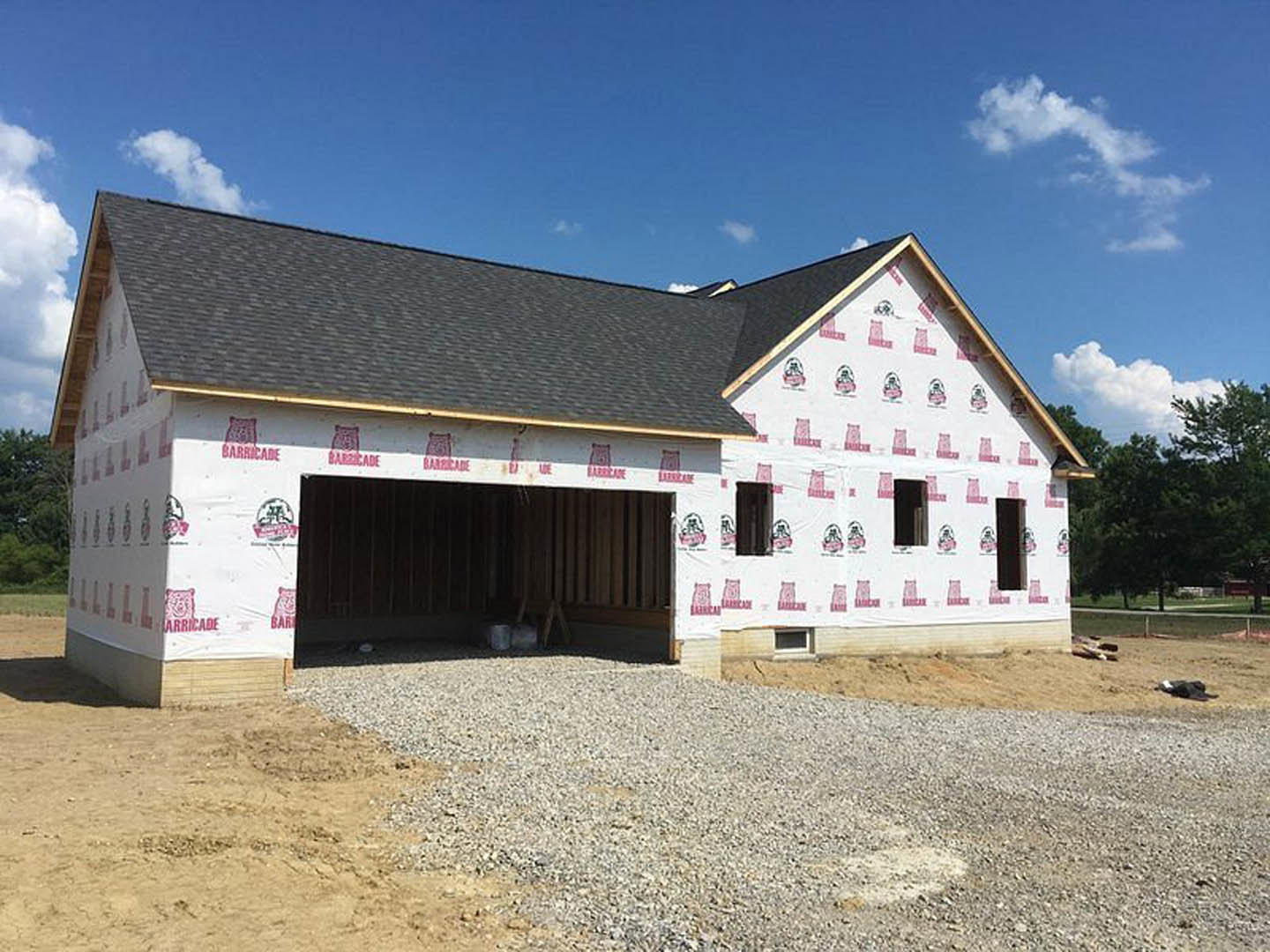 Framed custom home with attached garage, unfinished gravel driveway, exposed roof trusses, construction sign in foreground, wooded backdrop under cloudy sky