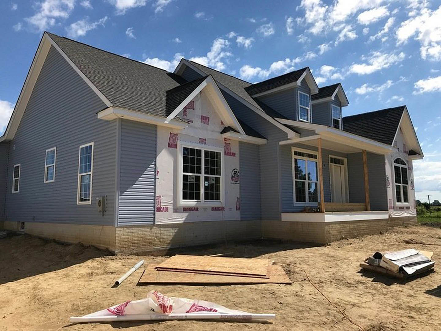 Partially built house with blue roof, exposed siding, and large window reflecting nearby tree; white and red construction bag and pile of newspapers scattered on dirt ground under