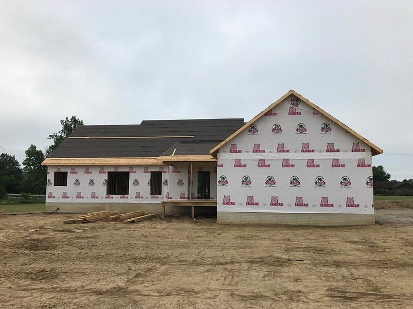 Framed house under construction with white roof covering, red and white stickers on exterior walls, workers on site, dirt lot, and temporary sign at the edge of the property