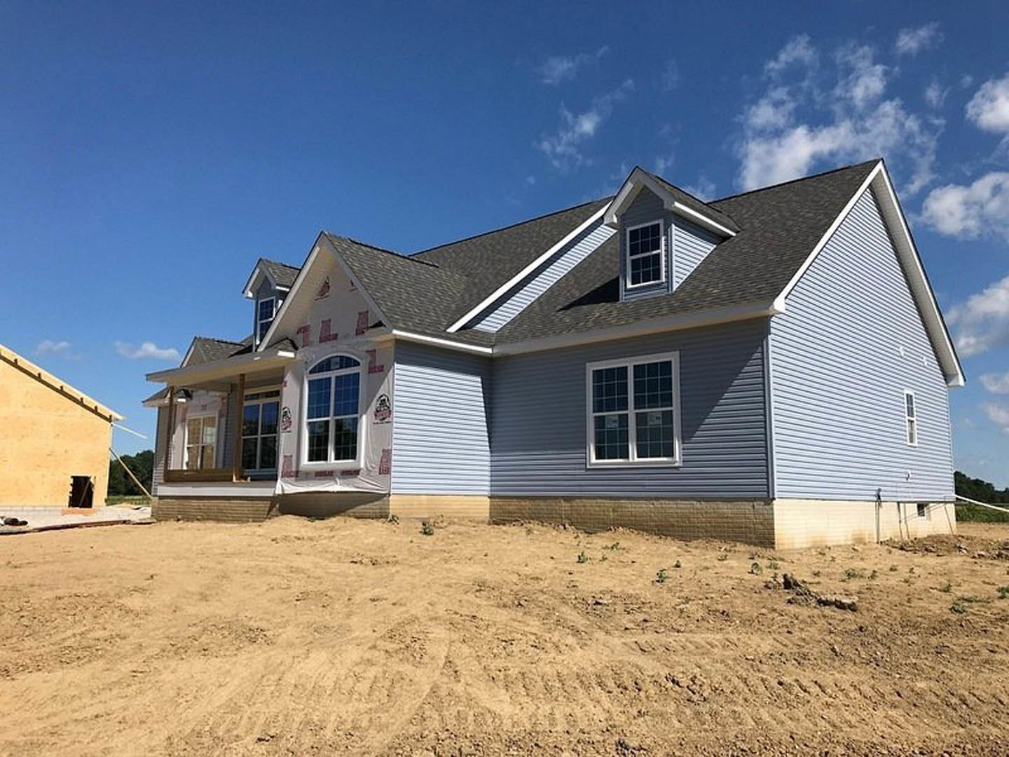 Two-story house under construction with blue roof, white-framed windows, open door, and exposed dirt yard under partly cloudy sky