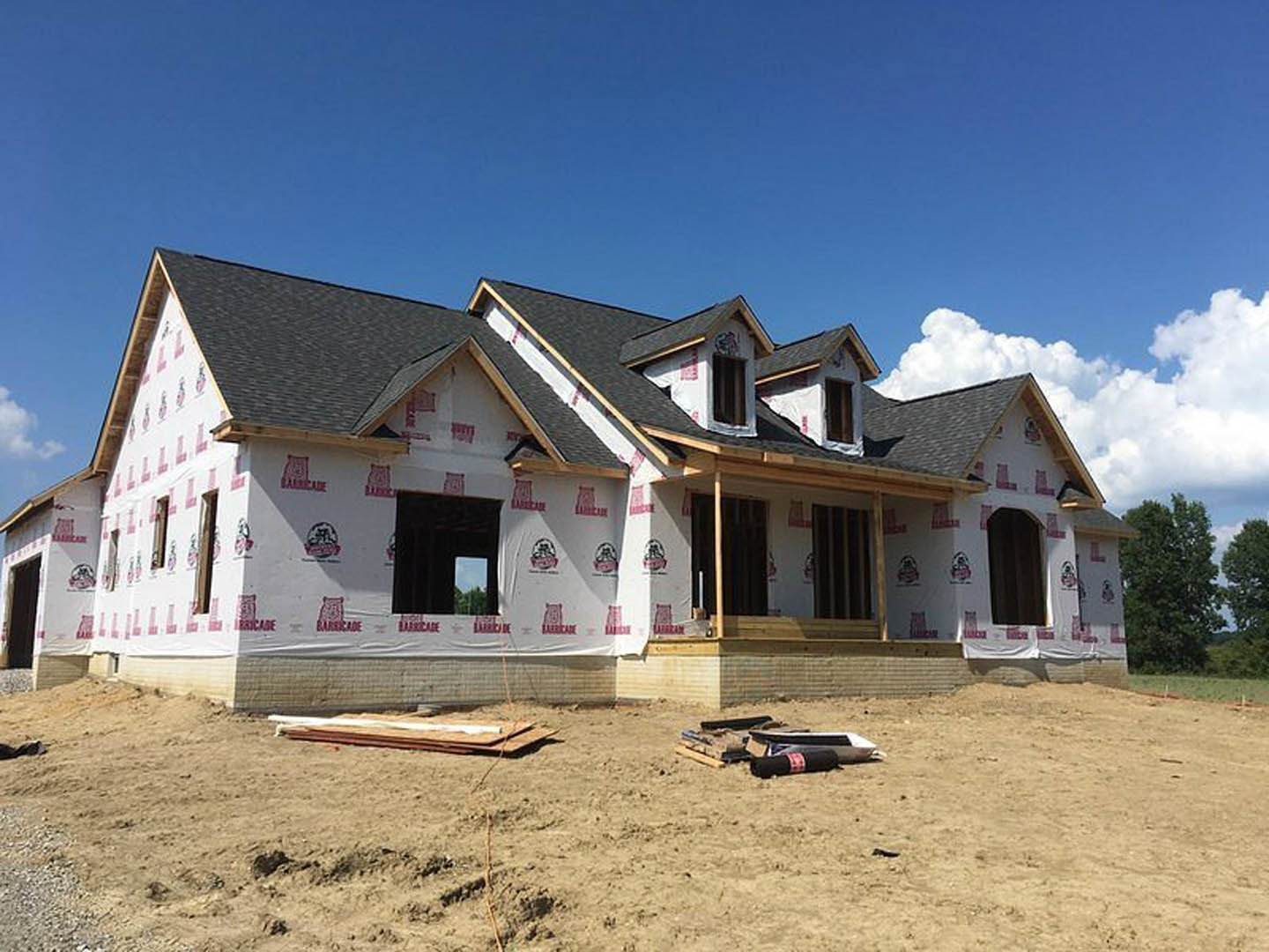 Partially built house with exposed framing, black doorway, window, red and white construction signs, dirt field scattered with pallets and wooden boards, leafy tree, wall