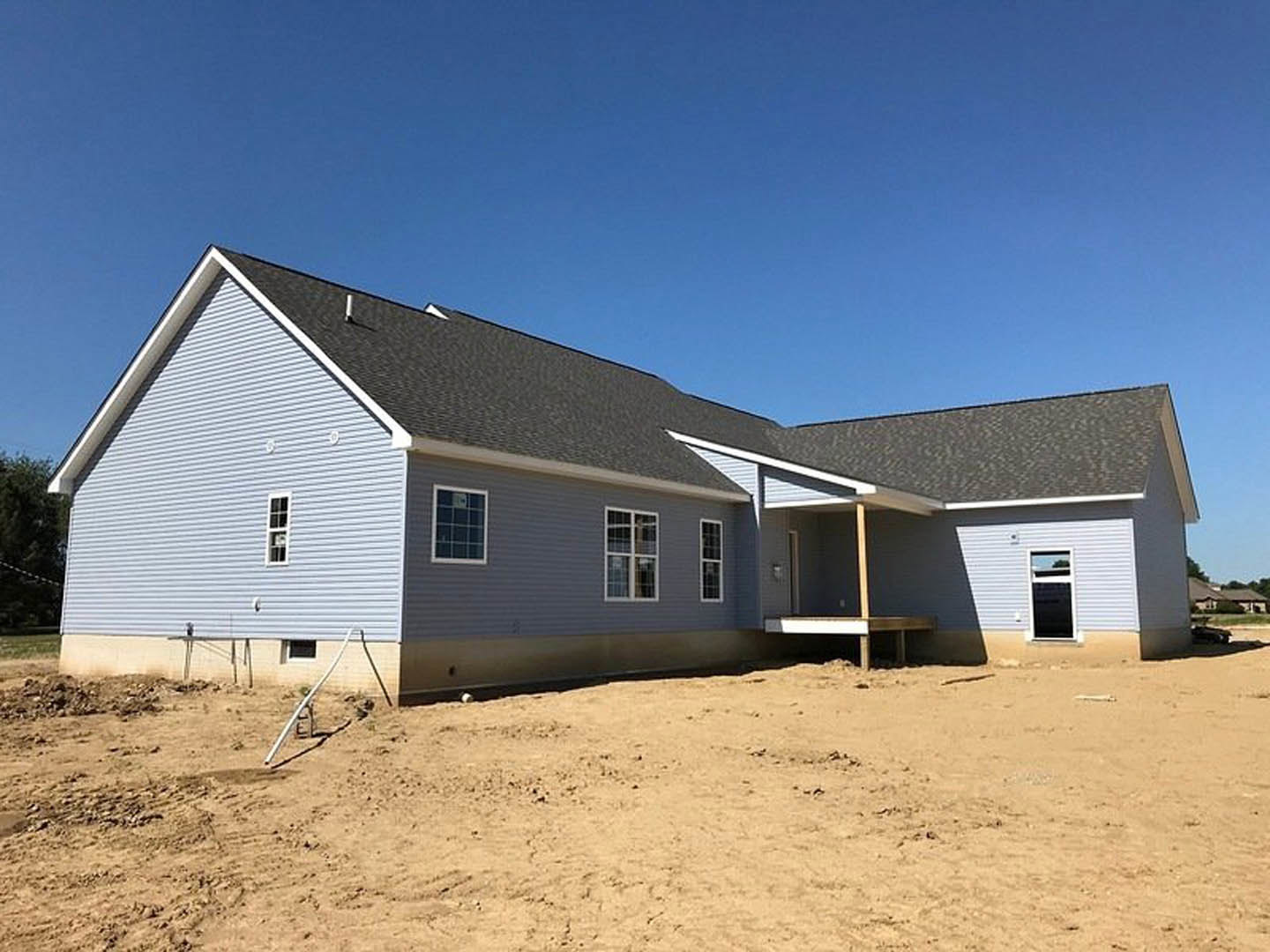 Wood-framed house under construction on sandy ground with exposed siding, windows, and clear blue sky