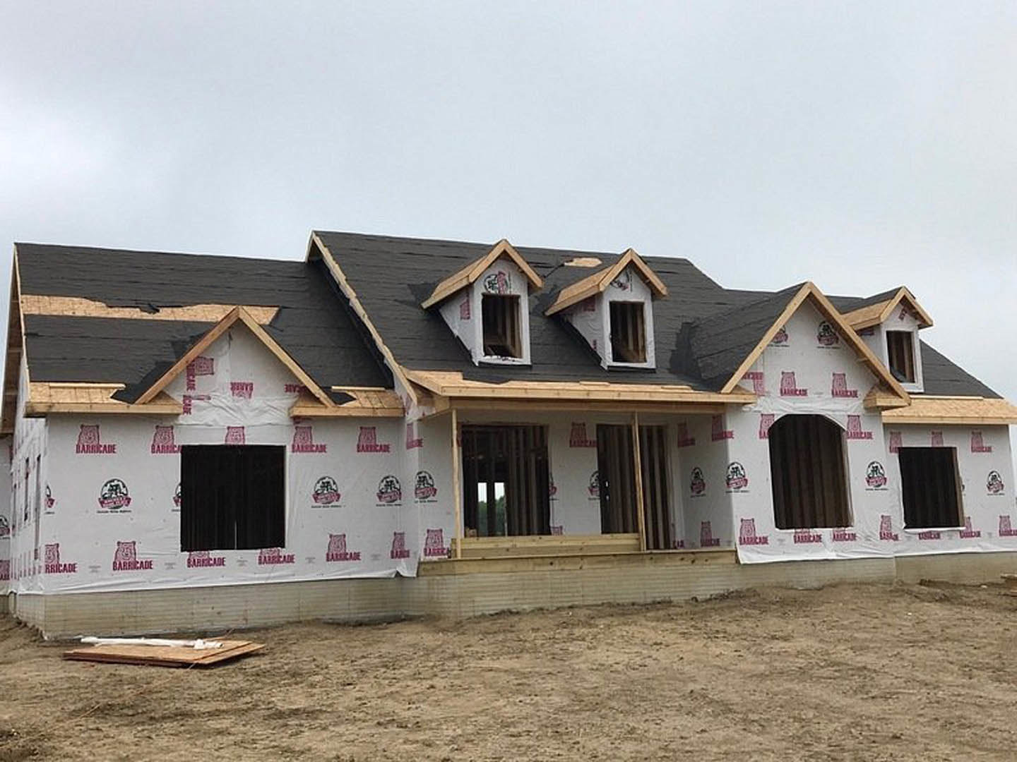 Two-story house under construction with black roof, exposed framing, and patch of dirt in front; large windows and unfinished exterior walls visible