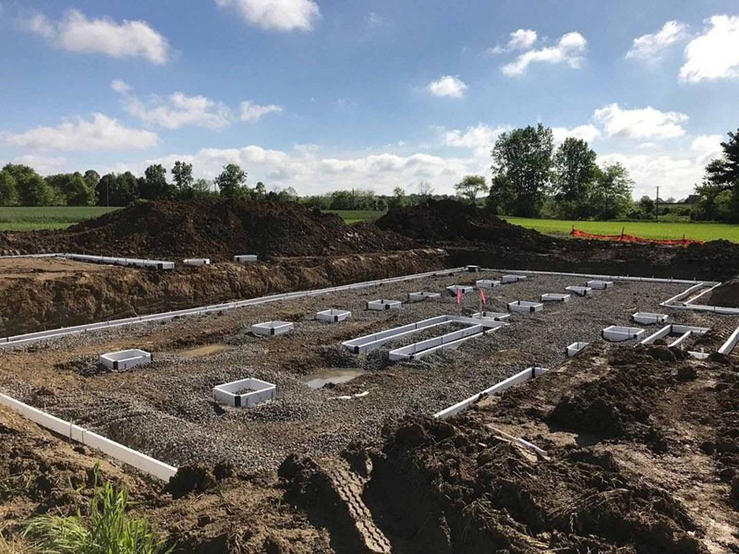 Dirt construction site bordered by trees, scattered concrete blocks, red pole, blue sky with clouds, white utility box filled with rocks, grassy field in foreground