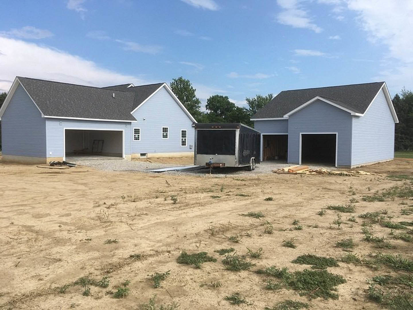 Modern single-story home with light siding, attached garages, and a black-covered trailer parked on a dirt driveway; cloudy sky overhead, open land surrounds property.