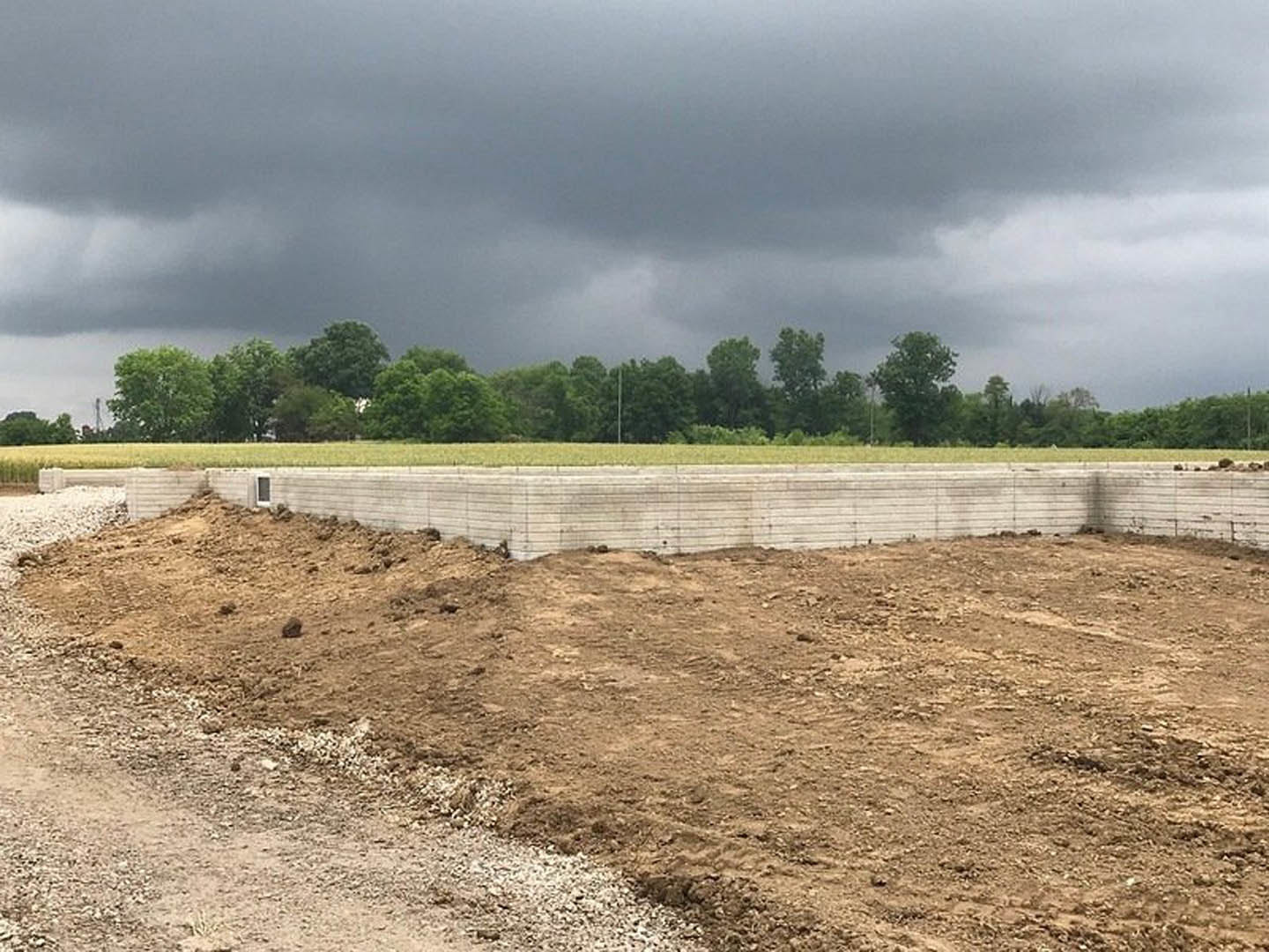 Dirt field with scattered rocks, concrete retaining wall, grassy area, and mature trees under a cloudy sky