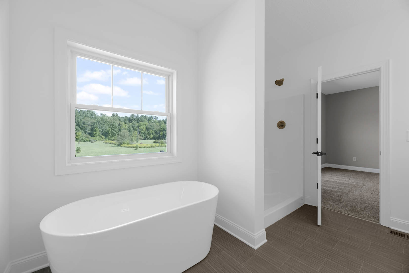 Bathroom with freestanding tub beneath large window, light-colored tile floor, white walls, and view of trees and field outside