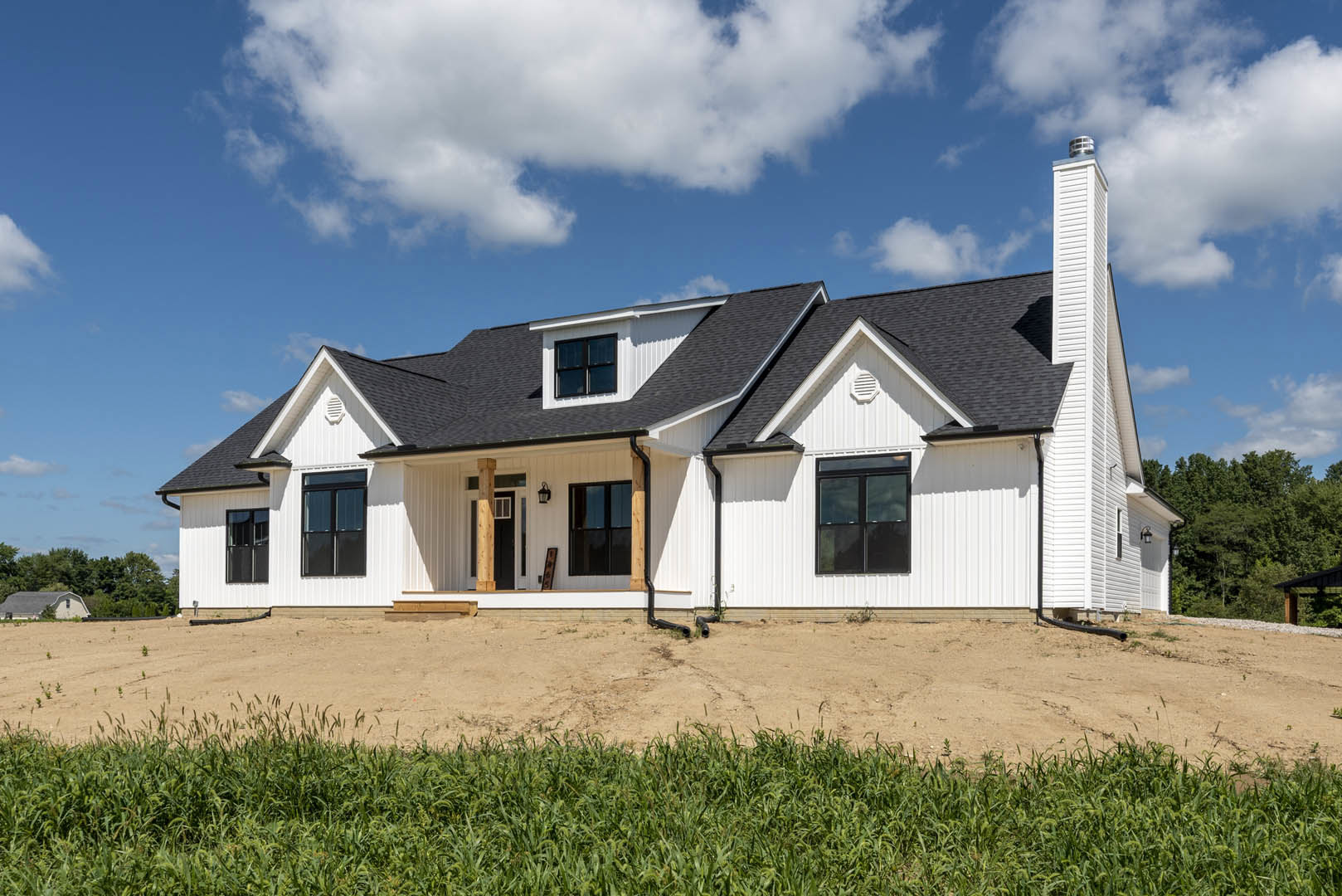 White house with black roof, large windows, and dirt field in foreground under blue sky with scattered clouds
