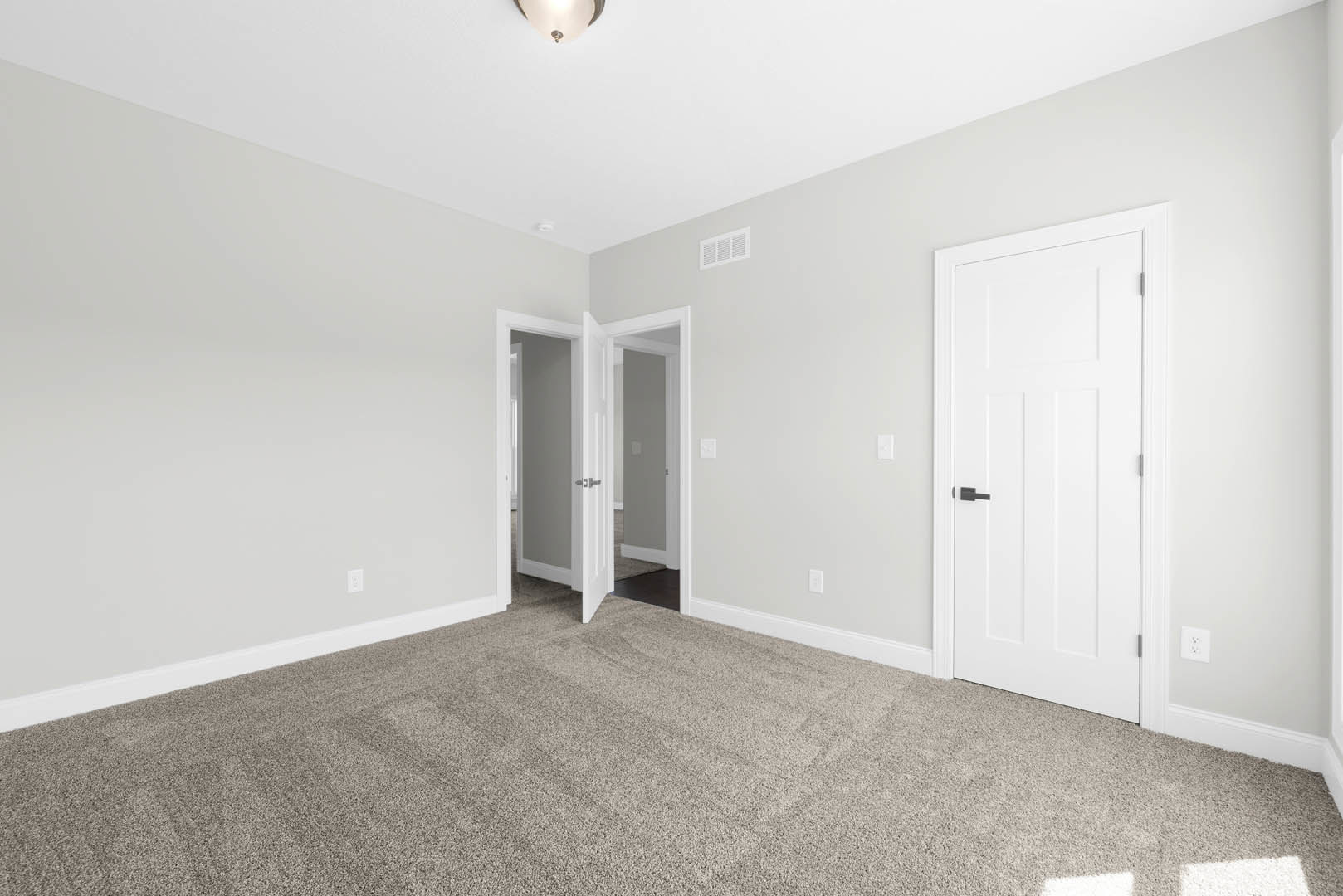 White-walled room with light carpet flooring, white door featuring black handle, silver door handles, and visible light switch on the wall