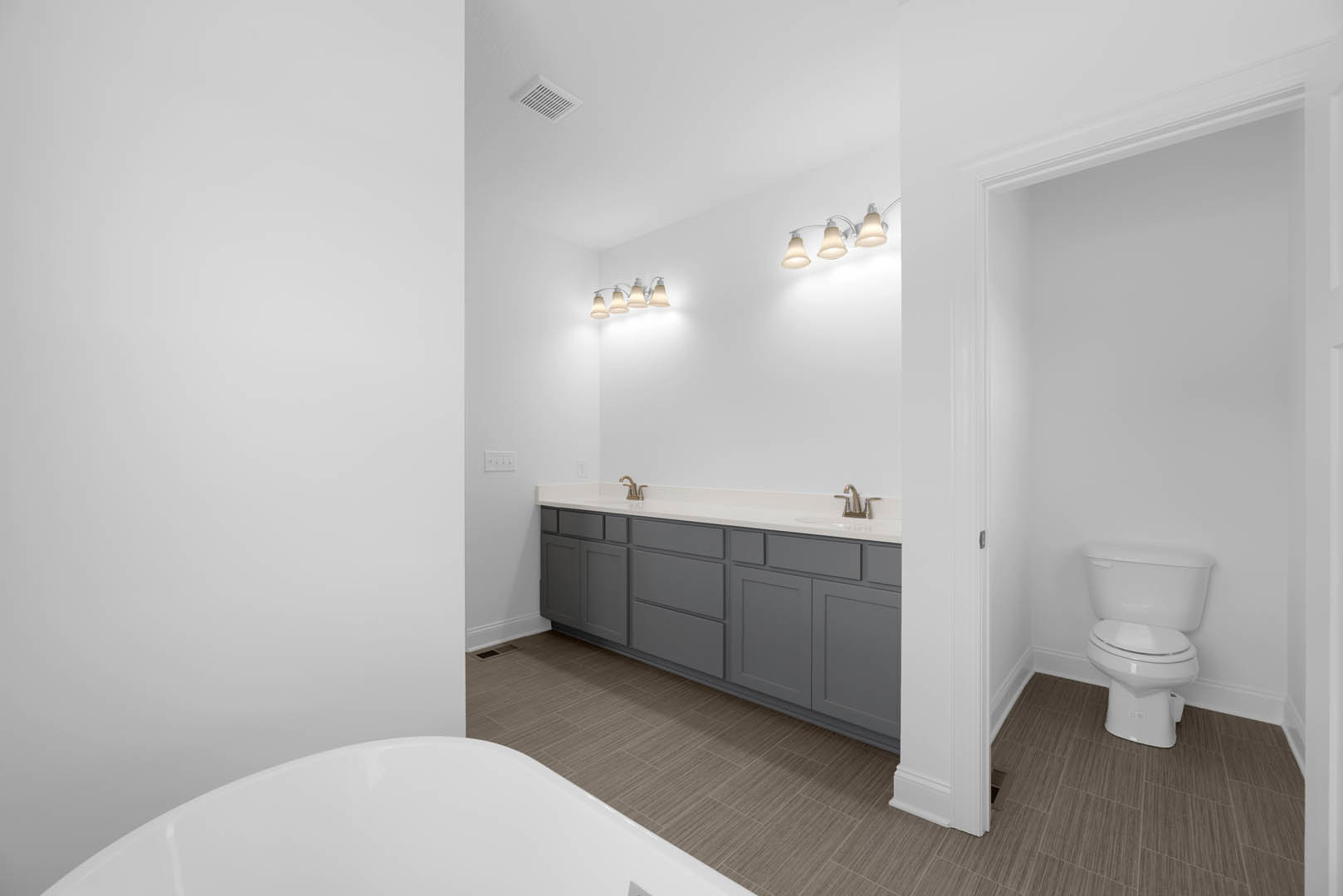 Bathroom with freestanding tub, white toilet, grey cabinets, wall-mounted sink, large mirror, and modern light fixtures above tile flooring