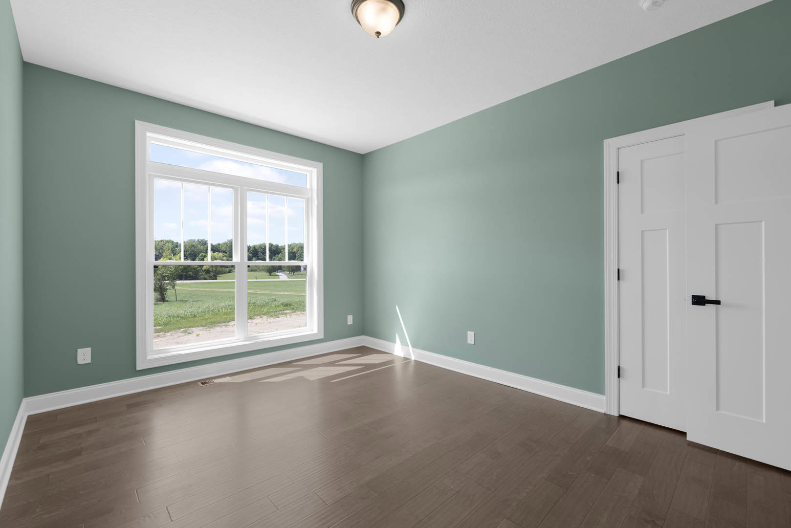 Bright room with laminate wood flooring, white walls, ceiling light fixture, large window overlooking grassy field, and door with black handle and decorative molding