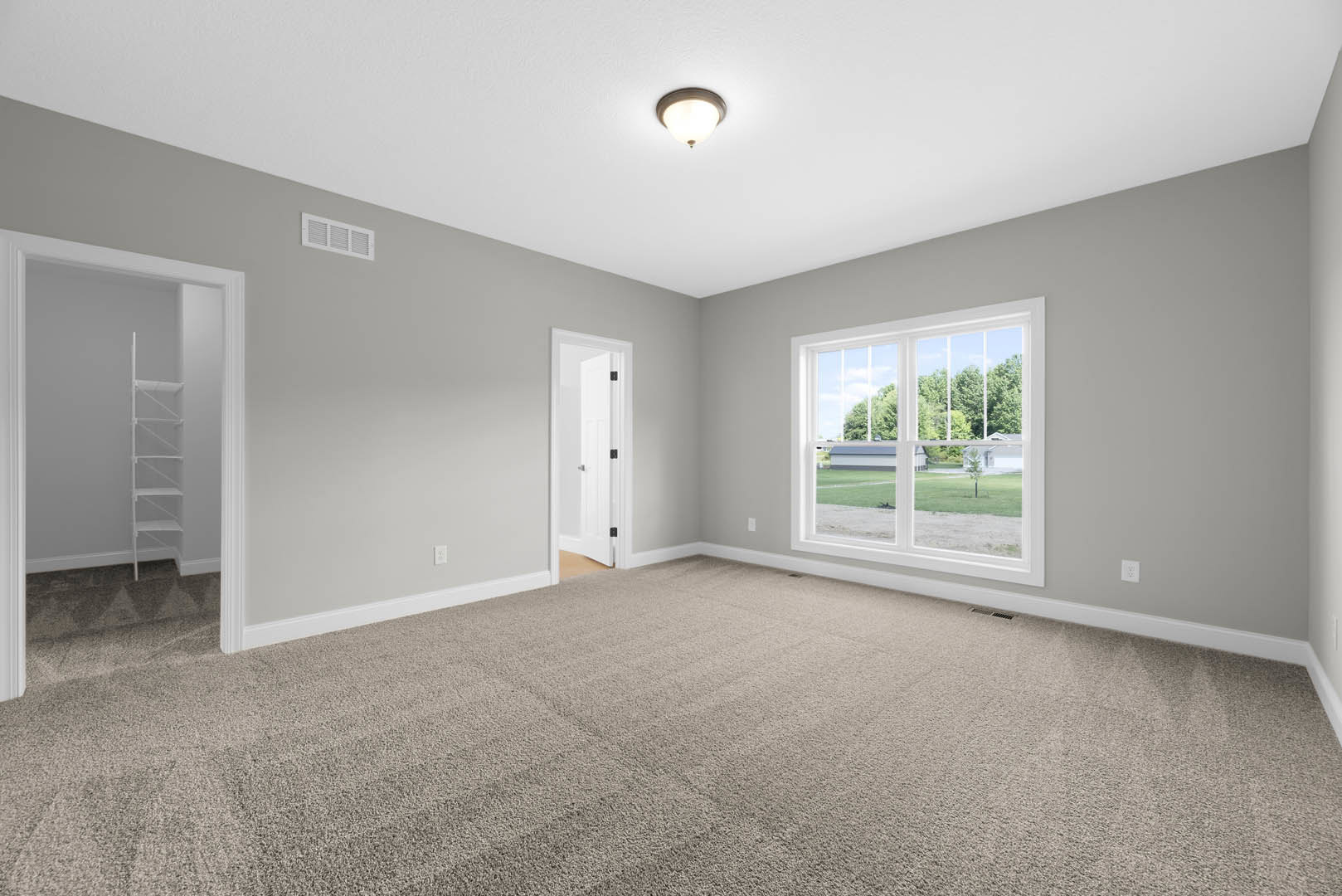 Neutral-toned carpeted room featuring a large window overlooking a green yard, white door with black handle, ceiling light fixture, and a white wall-mounted shelf.