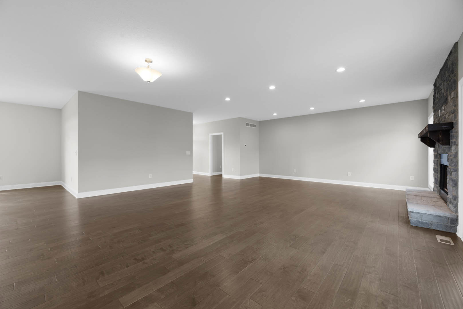 Hardwood floor in a spacious room with white plaster walls, stone fireplace set against a brick accent wall, and modern light fixture suspended from the ceiling