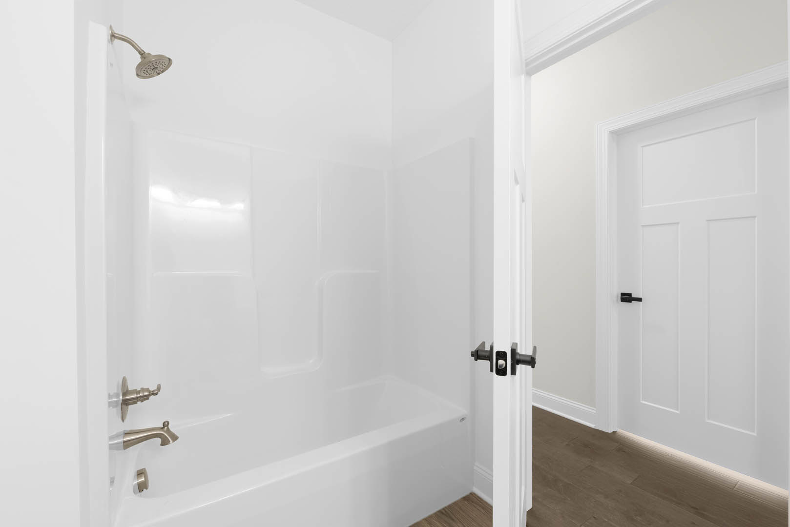 White bathtub and tiled shower with chrome faucet, black door handle on white door, light plaster walls, and ceiling.