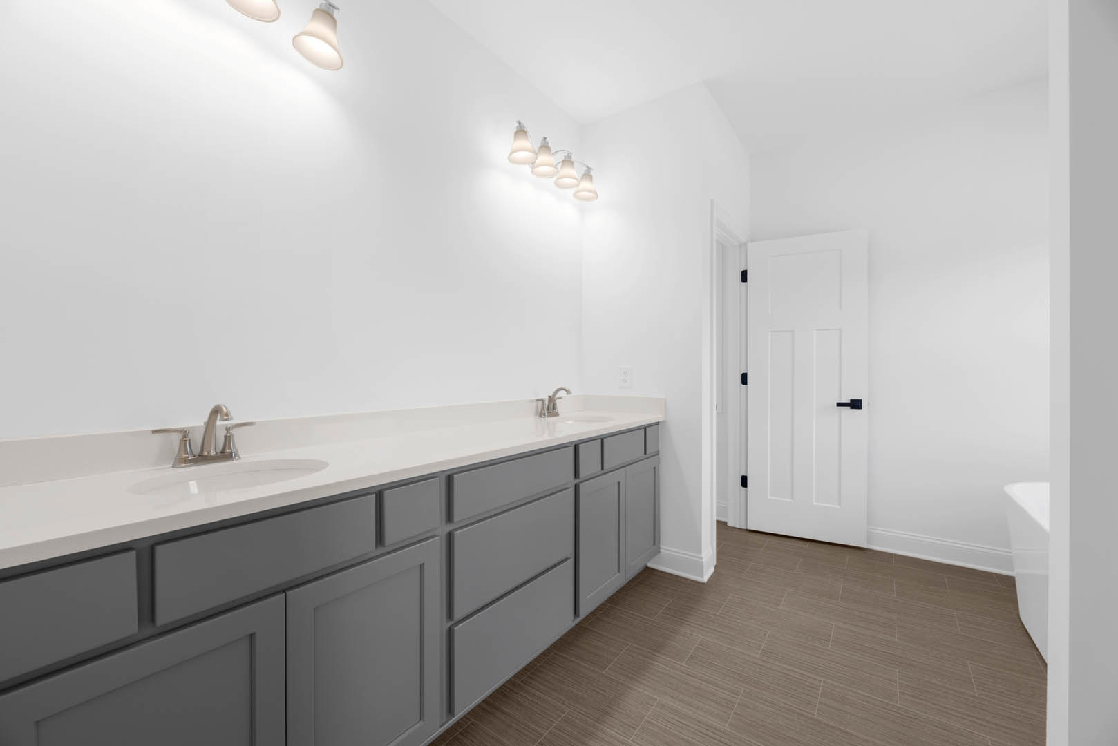 Double vanity with chrome faucets, white cabinetry, brown tile floor with white trim, white door featuring black handle, row of lights mounted on white wall