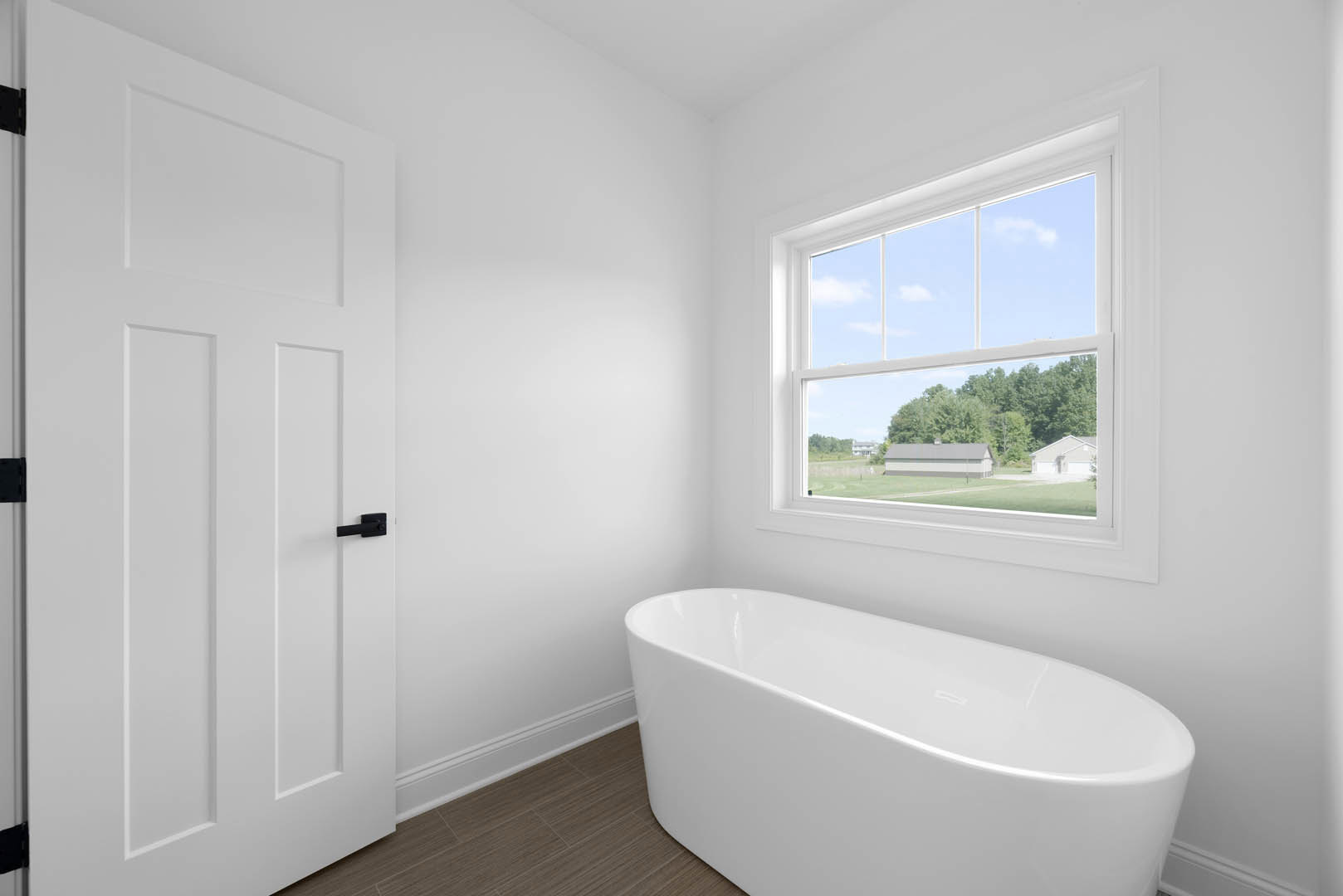 White bathroom featuring a freestanding curved bathtub beneath a window overlooking a field and trees, with light walls and chrome plumbing fixtures.