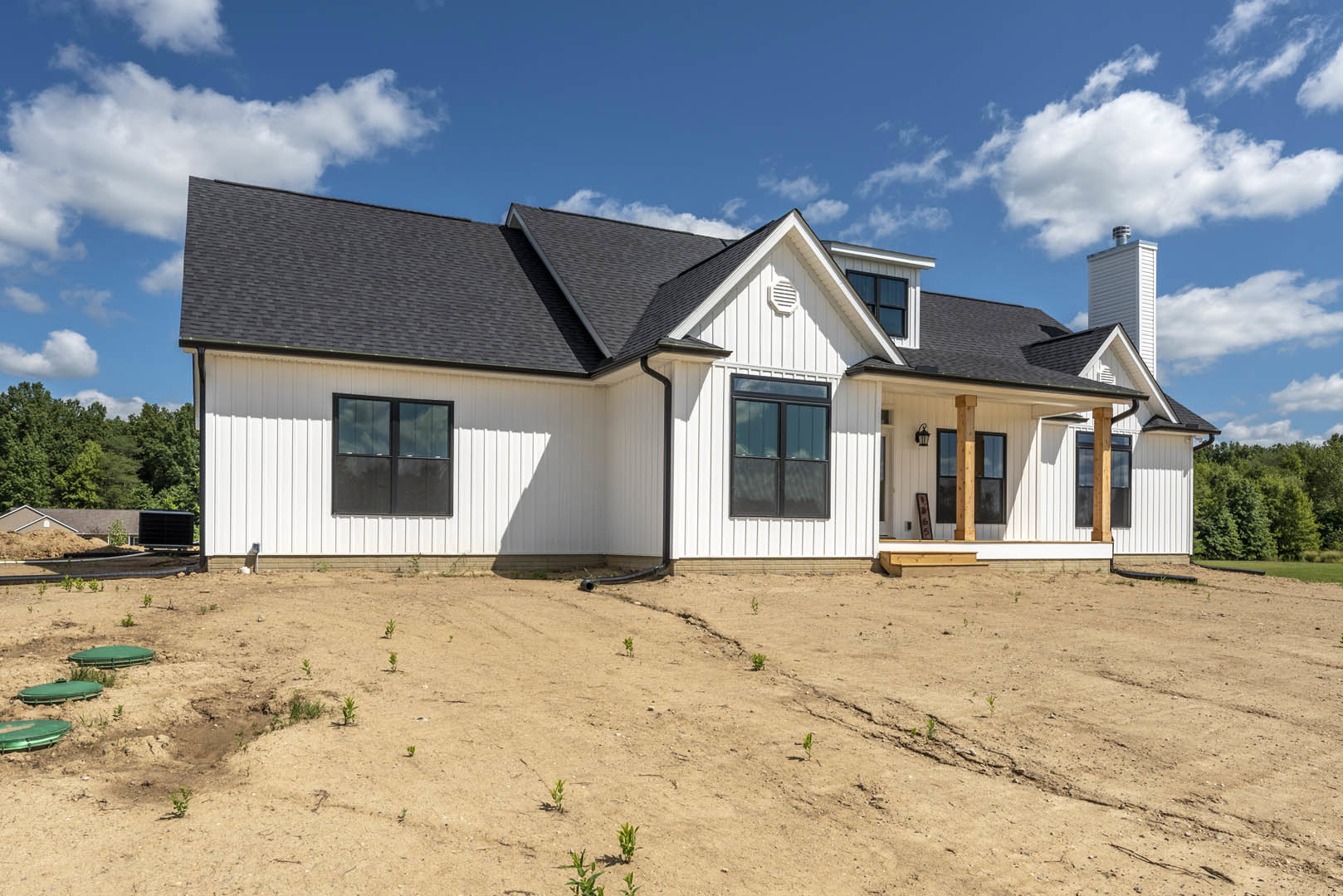 White house with black roof, large windows, dirt yard with small green plants, blue sky and scattered clouds