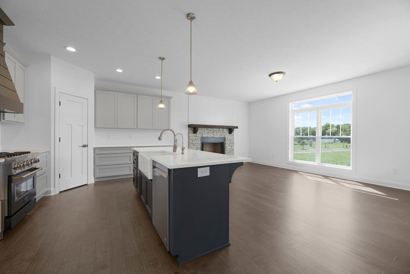 Kitchen with white countertop island, built-in fireplace, large window overlooking lawn and trees, stainless steel stove, white cabinetry, black hardware, illuminated pendant light