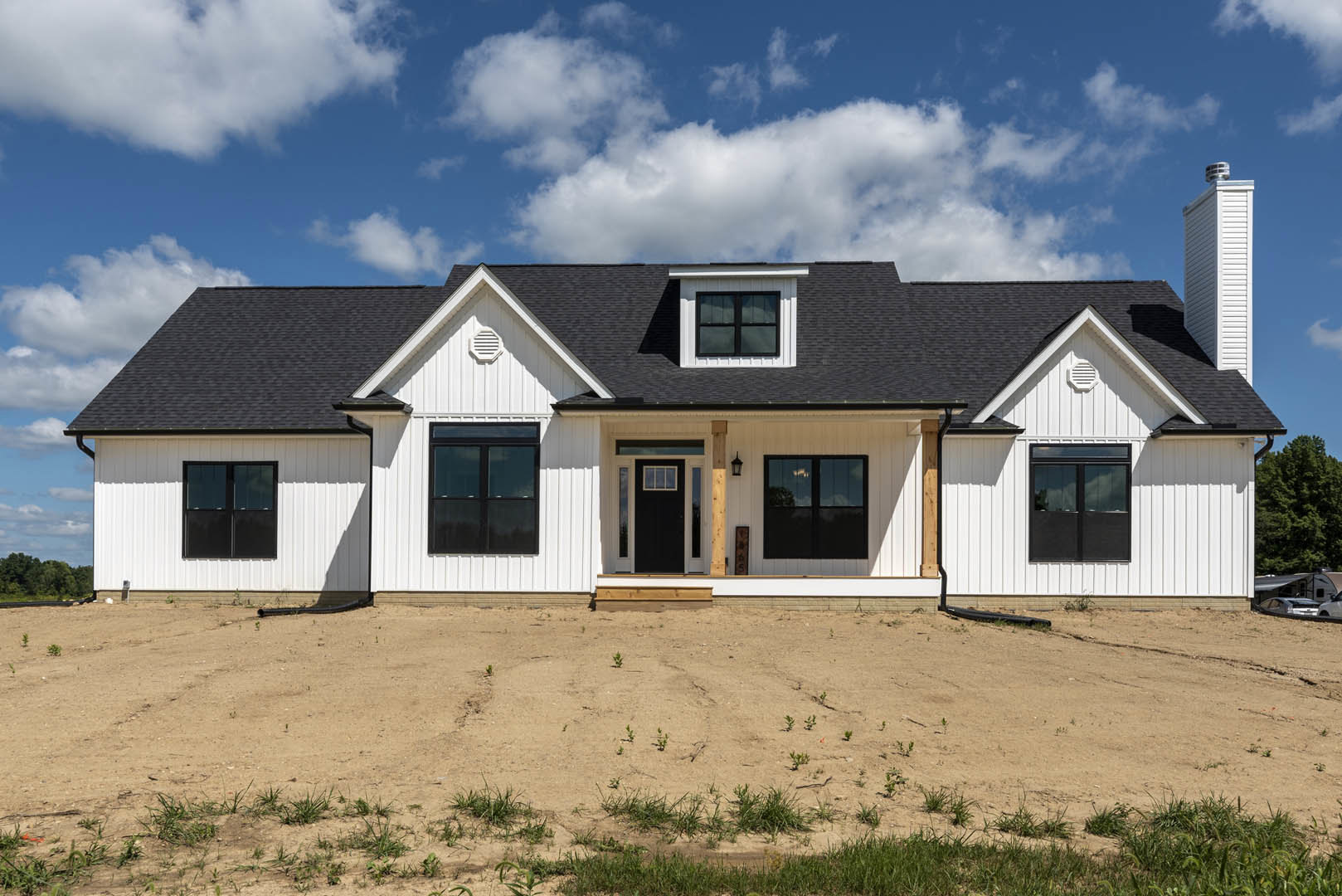 White siding house with black-framed windows and double black front doors reflecting a tree, dirt yard with sparse small plants, cloudy sky overhead