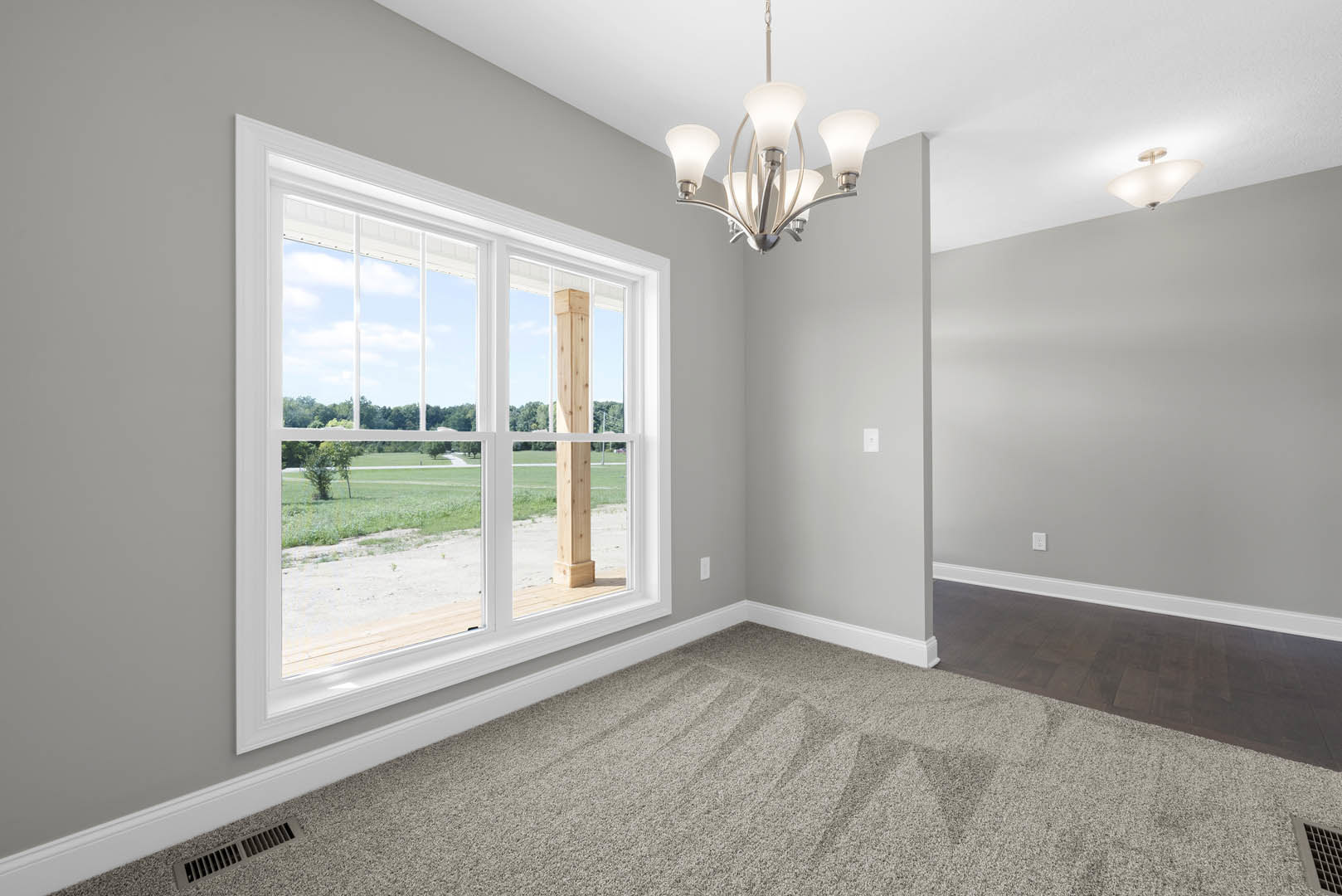Carpeted room with large window overlooking grassy field, white walls with crown molding, wooden post, chandelier, and floor vent