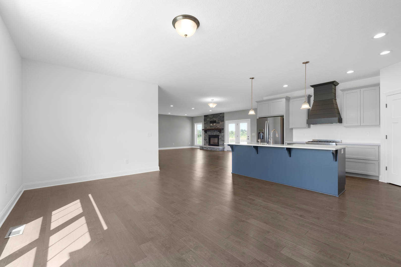 Open-concept kitchen and living room featuring wood flooring, blue and white kitchen counter, white cabinetry, stainless vent hood, modern light fixture, glass-front fireplace, and