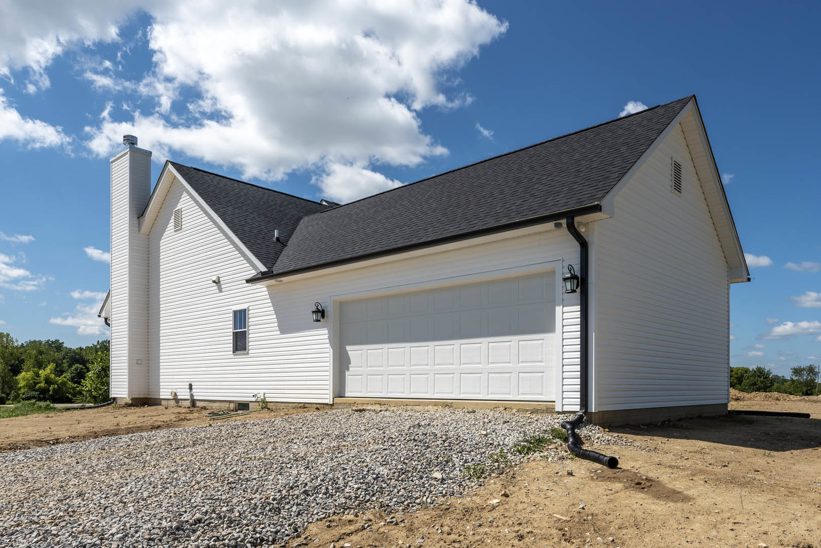 White siding house with attached garage, gravel driveway, exterior wall lights, black drainage pipe, pile of rocks, and surrounding forested trees under cloudy sky.