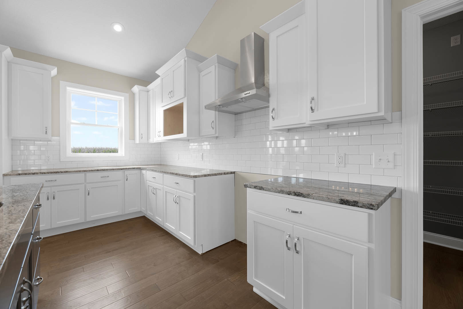White kitchen cabinets with marble countertops, wood flooring, stainless steel appliances, large window overlooking a field, undermount sink, and vent hood.