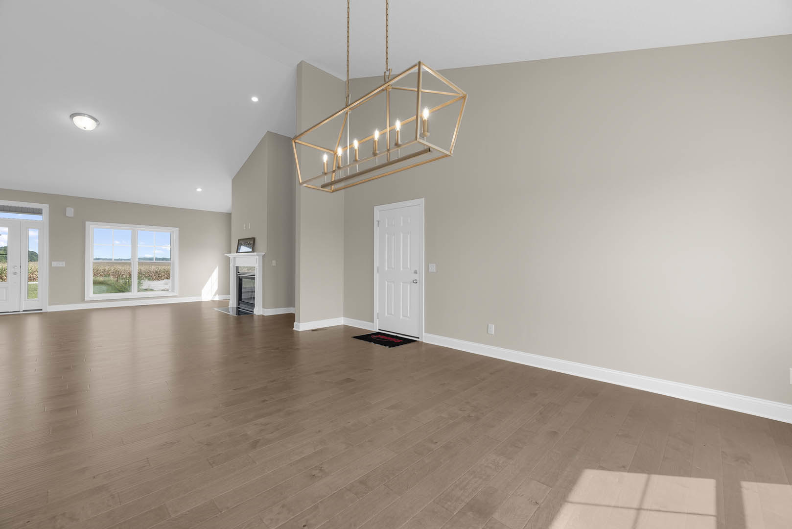 Chandelier hanging from white ceiling, wood flooring with patterned rug, white door with silver knobs, large window overlooking grassy field, plaster walls